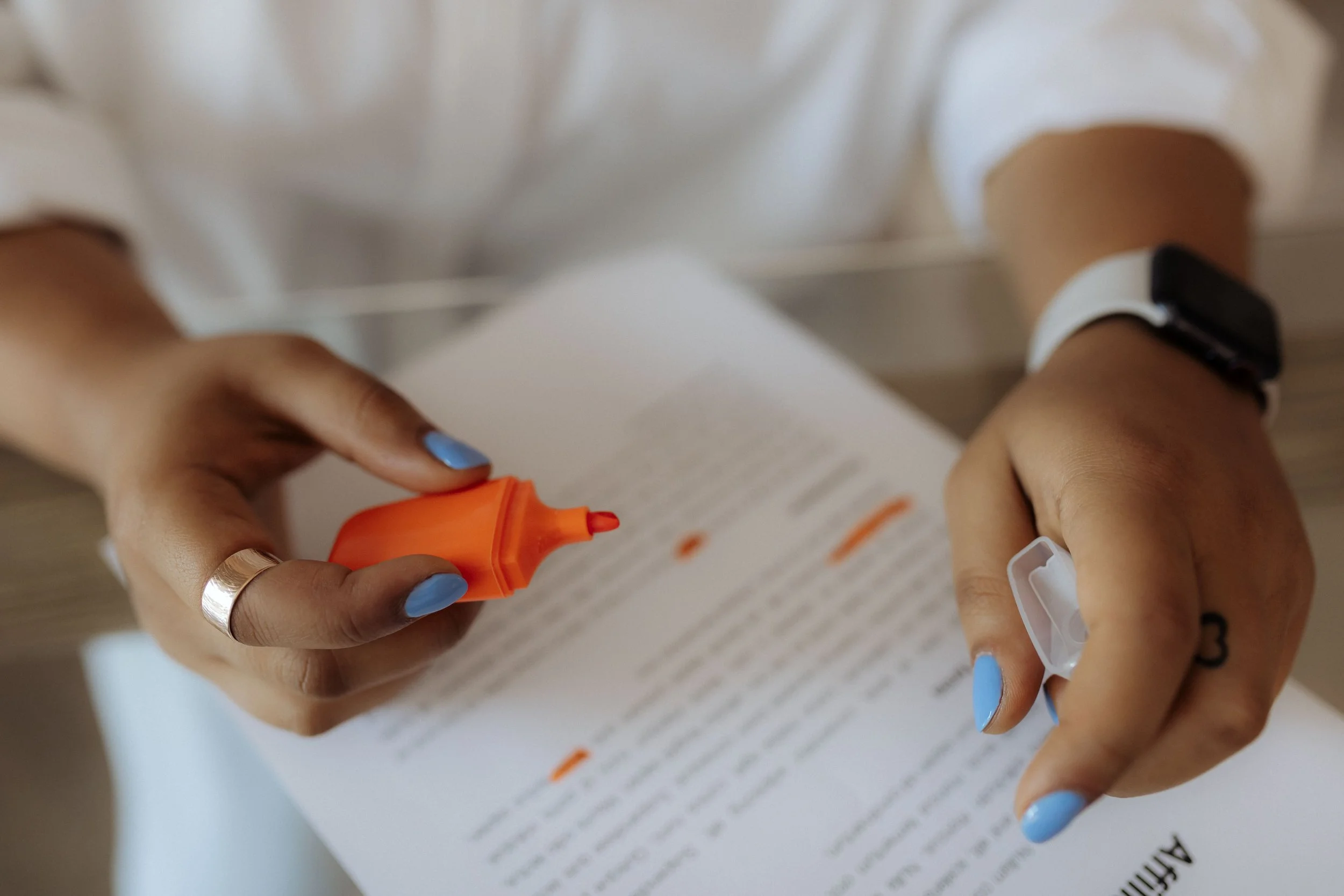 Person applying orange correction fluid to a document with blue nail polish and wearing a smartwatch.