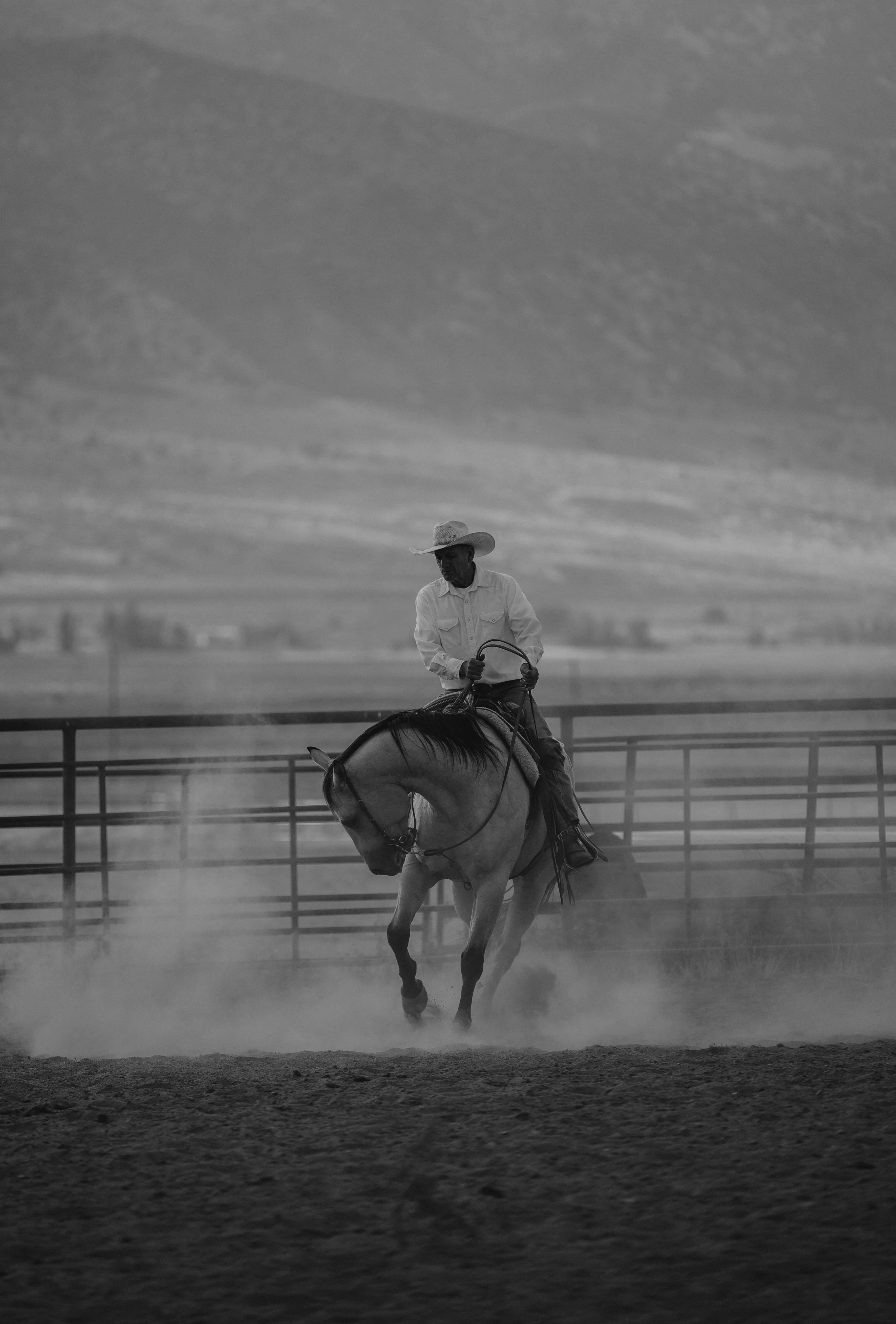 A man in cowboy attire riding a horse in an outdoor arena with dust clouds, countryside landscape in the background, black and white photo. Arena photoshoot. Western Photography.