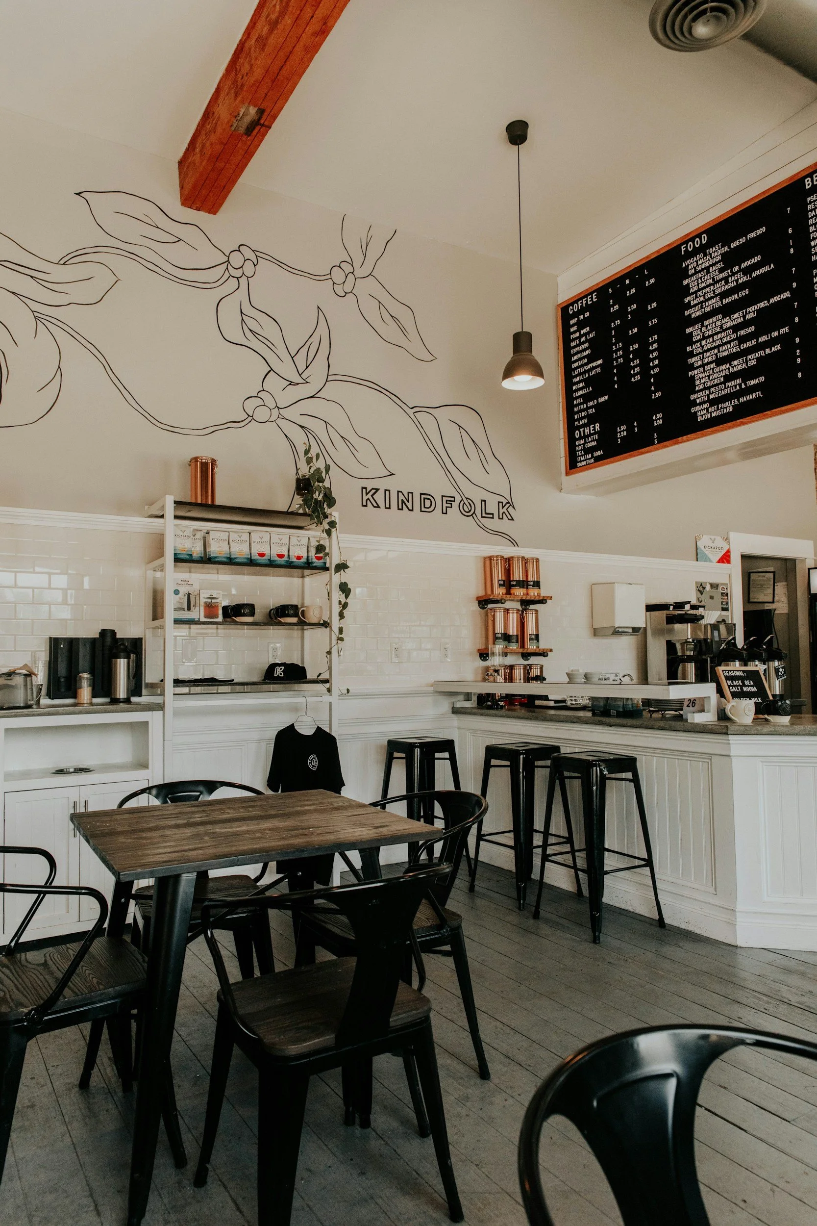 Interior of a modern cafe with black chairs, wooden tables, and a white counter. A large black menu board hangs on the wall, and a wall painting features a botanical line drawing with the word 'KINDFOLK'.