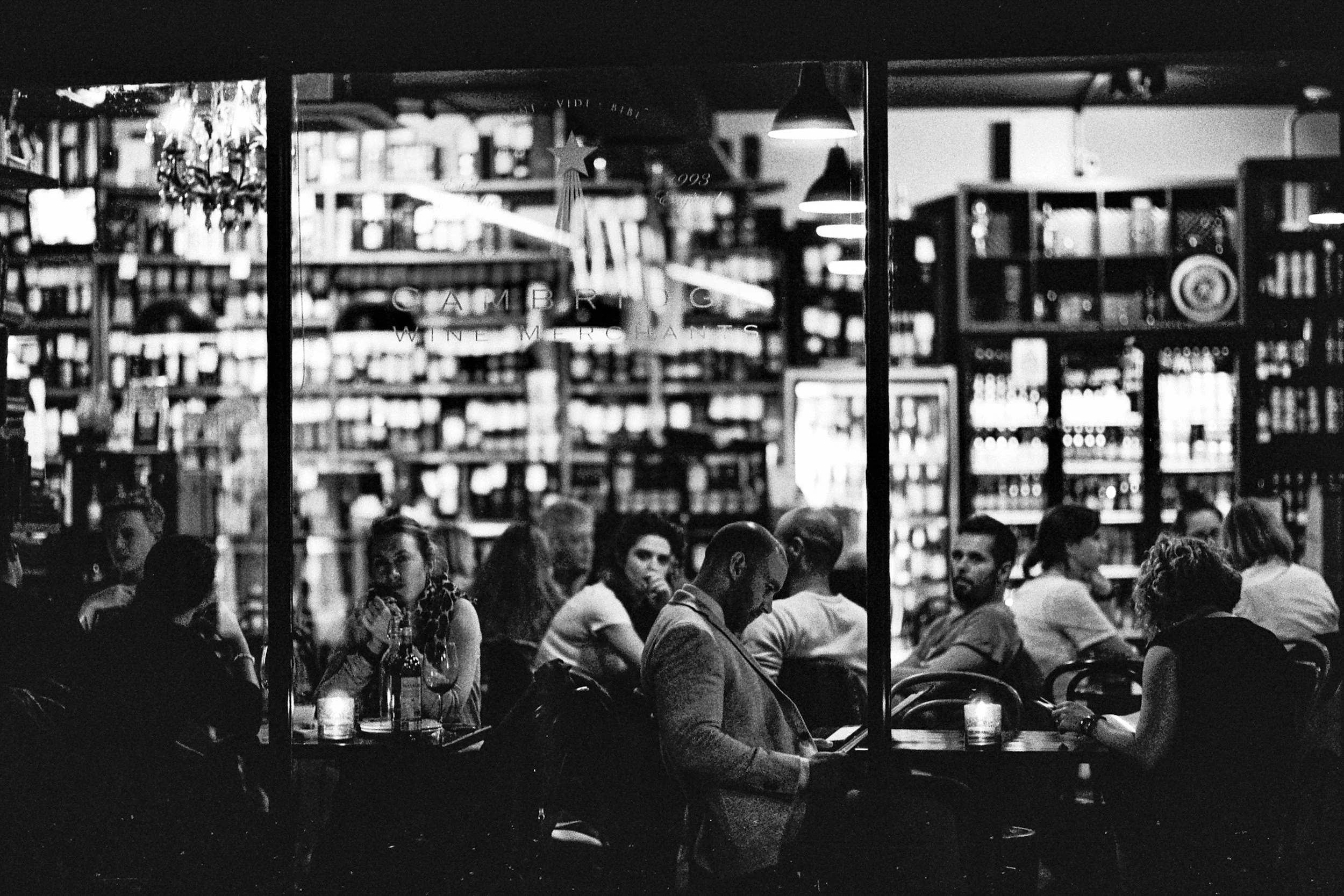 Black and white photo of people dining at a restaurant with shelves of bottles in the background and a window with a sign labeled 'CAMPBELL WINEMAKERS'