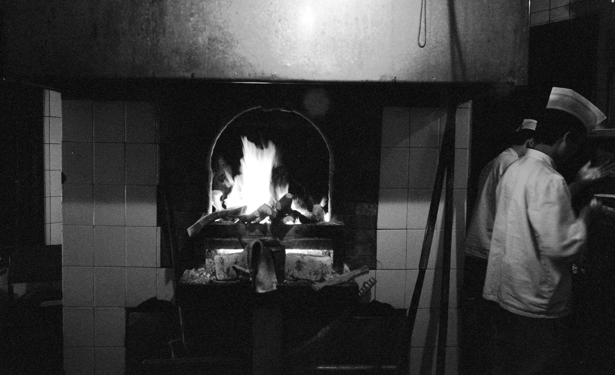 A black and white photo of a fireplace with a roaring fire. Two chefs, dressed in white uniforms and hats, stand to the right, preparing food in a kitchen.