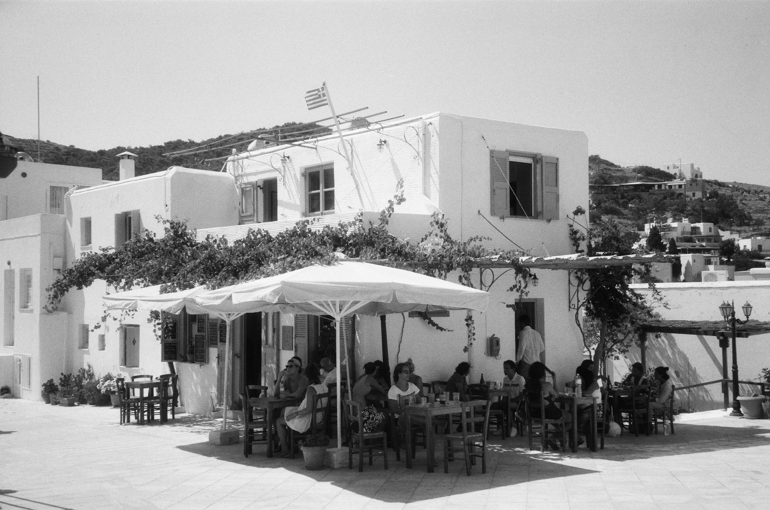 Outdoor café with tables and chairs under umbrellas, people sitting and dining, white buildings with shutters, hillside in the background, in a sunny setting.