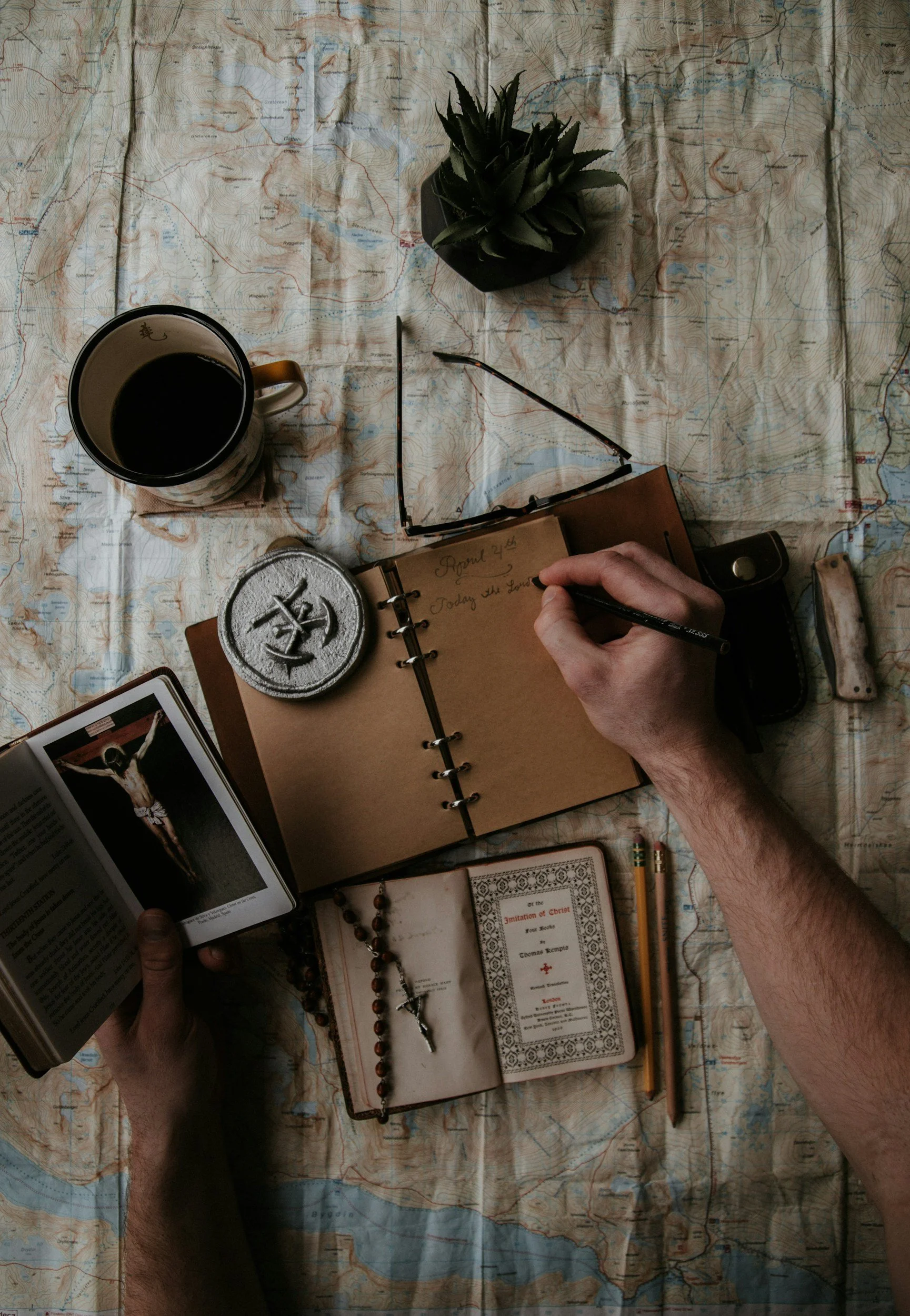 A person writing in a notebook on a map-covered surface, surrounded by various objects including a cup of coffee, a potted plant, a book, a rosary, a compass, and other small items.