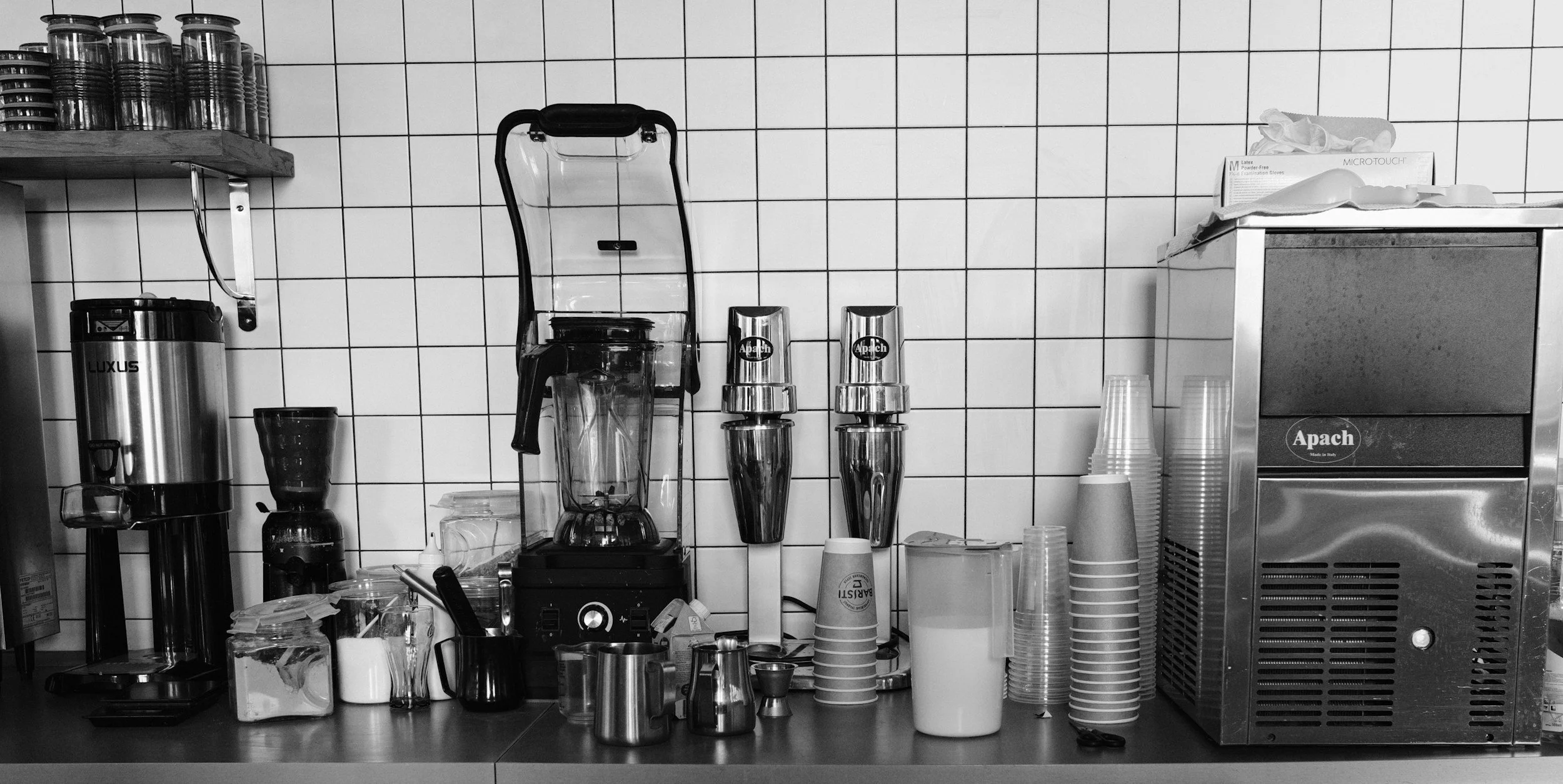 A black and white photo of a coffee shop or kitchen counter with various appliances and items, including blenders, paper cups, and containers, against a tiled wall.