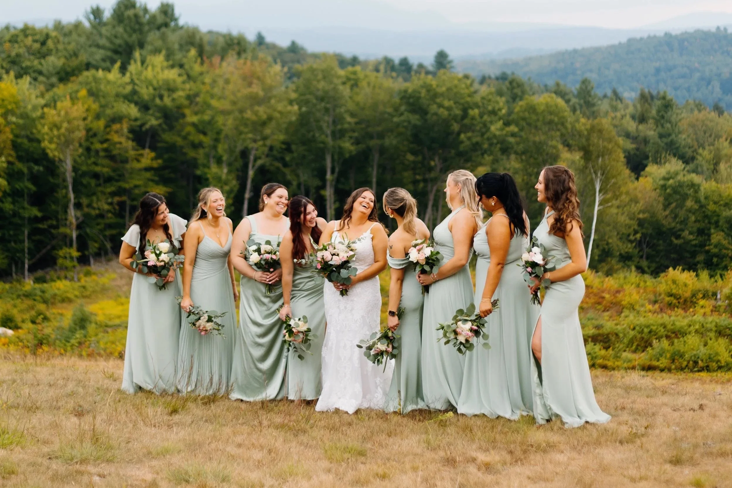 A group of women in bridesmaid dresses and one bride in a white wedding gown, standing outdoors in a grassy field with trees and mountains in the background, smiling and holding bouquets of flowers.