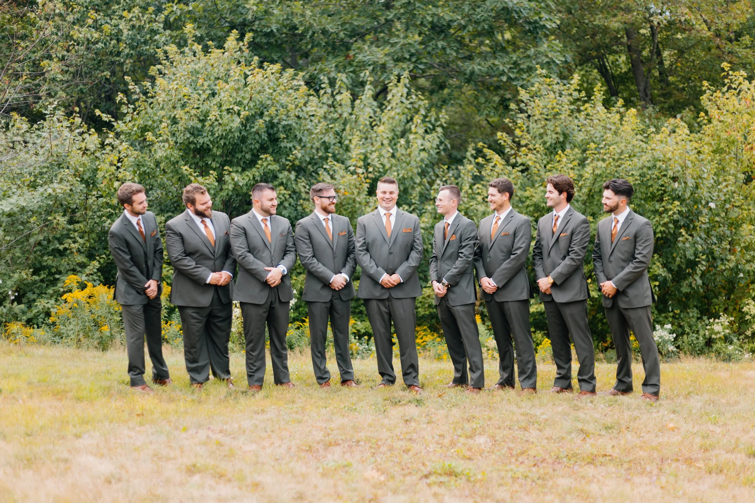 Group of nine men dressed in matching gray suits with orange ties and pocket squares standing outdoors on a grassy area with green trees in the background, smiling and looking at each other.