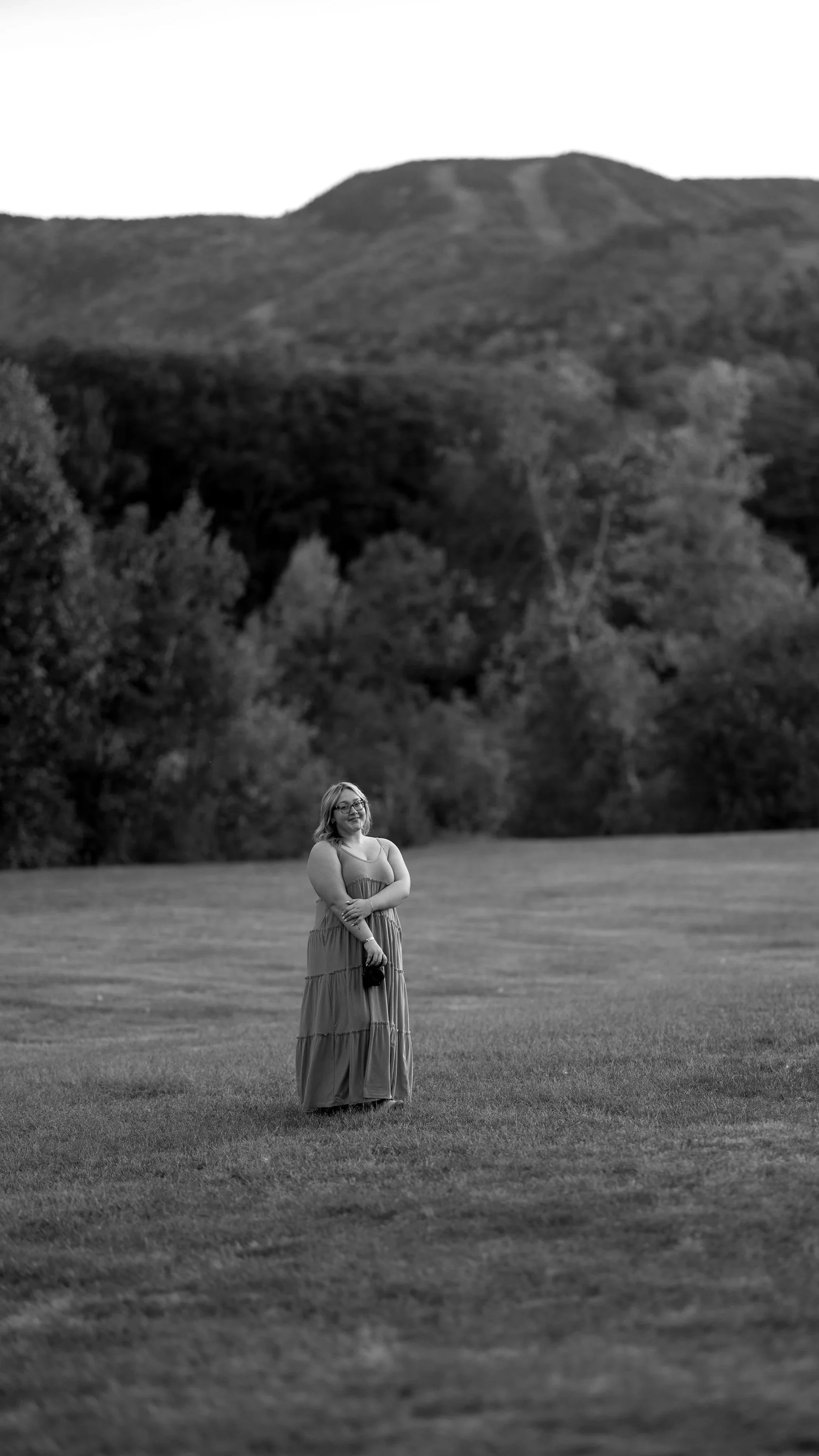 A woman in a long dress holding a camera stands on a grassy field with a backdrop of trees and mountains in black and white.