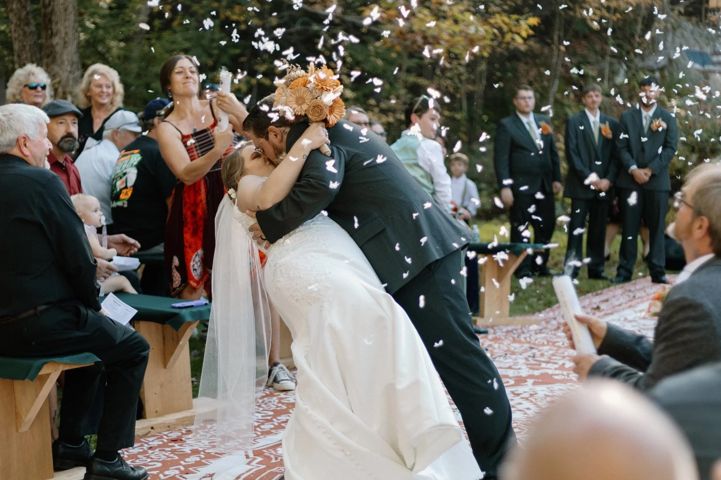 A newlywed couple sharing a kiss at their outdoor wedding ceremony, surrounded by friends and family, with guests smiling and taking photos as confetti falls.