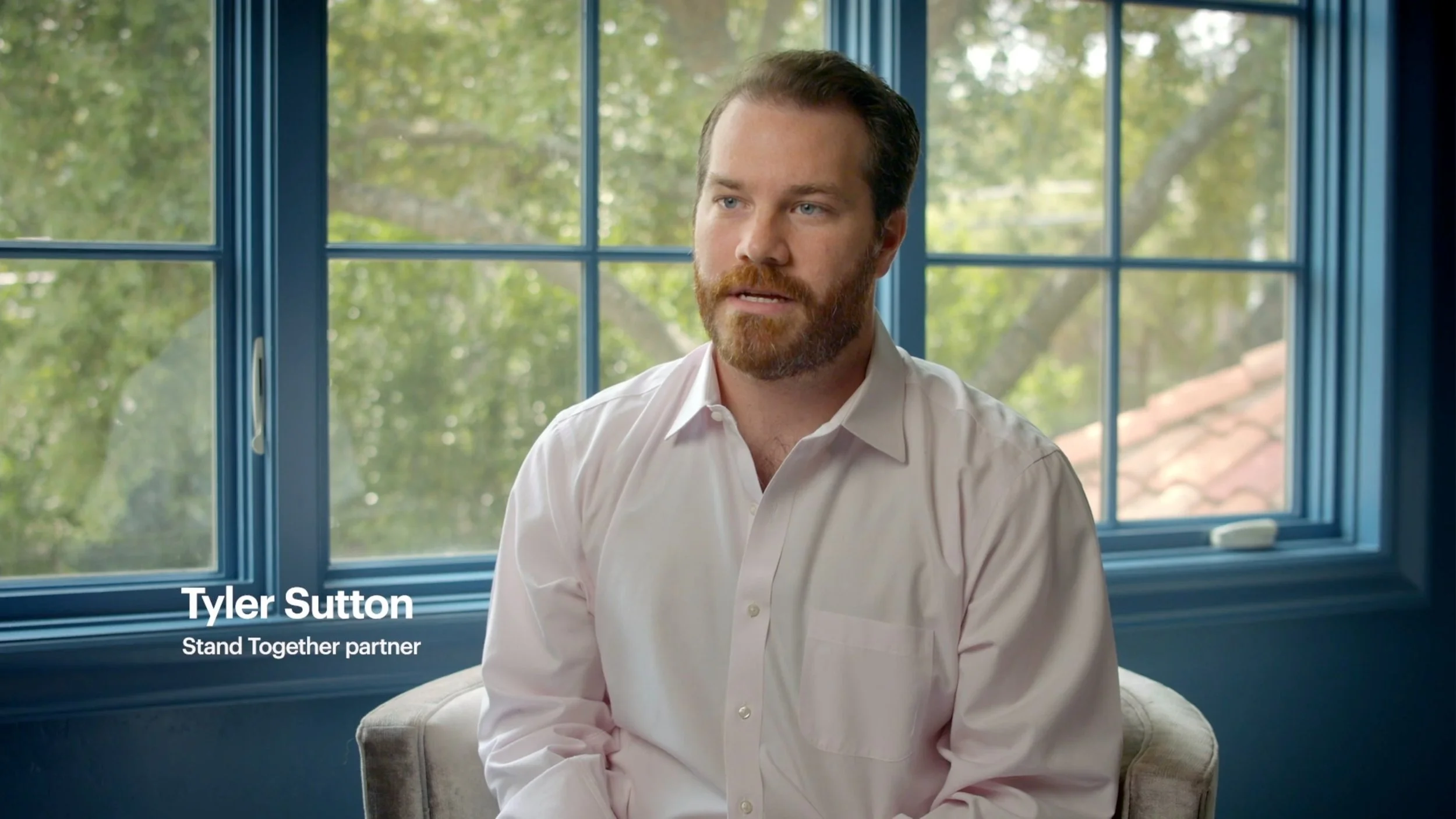 A man with a beard and mustache, wearing a white dress shirt, sitting indoors near a window with blue frame, with trees and a tiled roof visible outside.