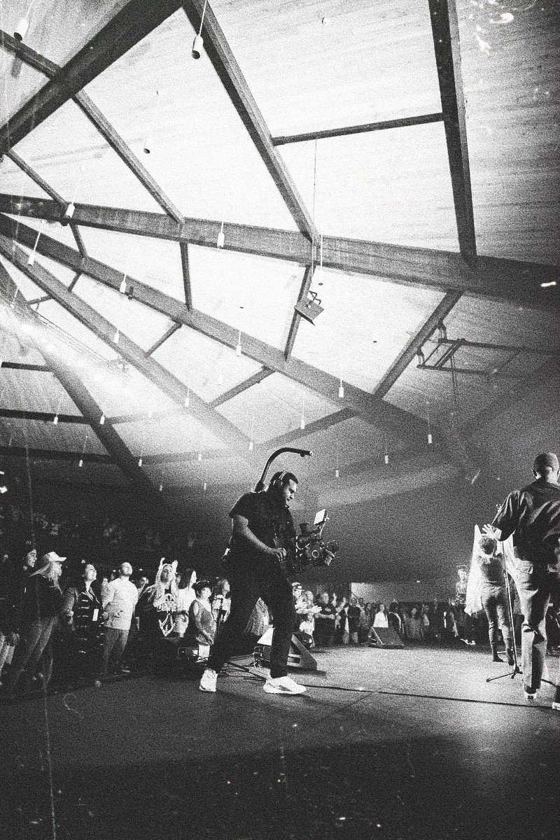 A crew member with headphones and equipment operating a camera on a stage with a crowd watching under a large ceiling with skylights.
