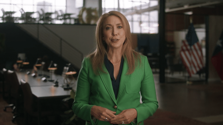 A woman with blonde hair, wearing a green blazer and black blouse, speaking in a modern room with a glass wall, American flags, and a desk with computers.