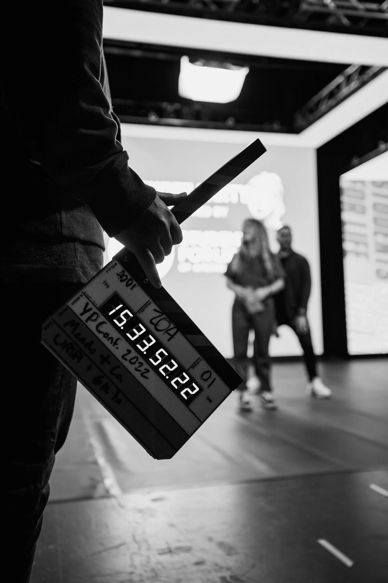 A person holding a film slate on a film set with two people in the background, one holding a script, in an indoor studio with stage lighting.