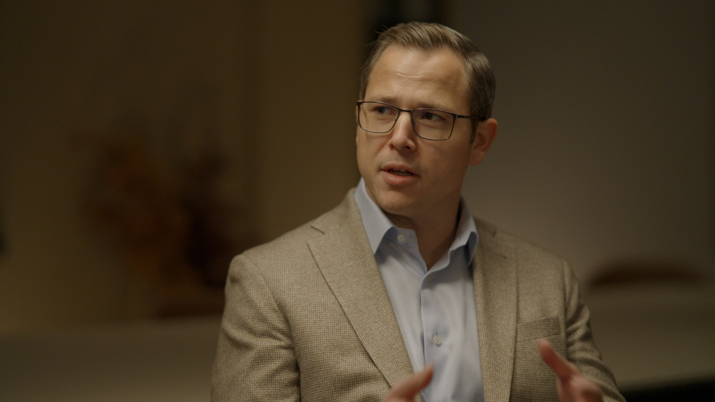 A man with glasses and neatly combed hair, wearing a light brown blazer and white collared shirt, appears to be speaking or explaining something in an indoor setting with a blurred background.