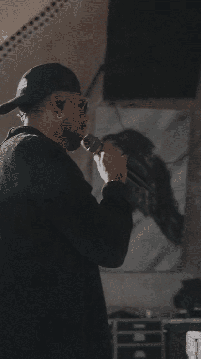 A person wearing a black cap, glasses, and earrings, holding a microphone and speaking, with long hair and a beard, indoors.