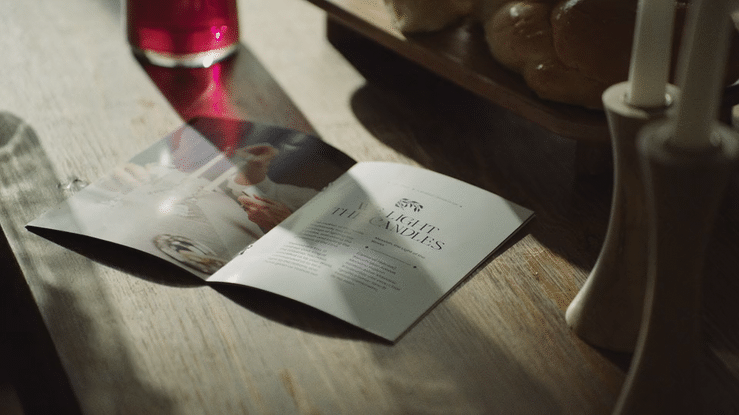 Open magazine with text and images on a wooden table next to a red glass, a bread basket, and a white ceramic holder with candles.