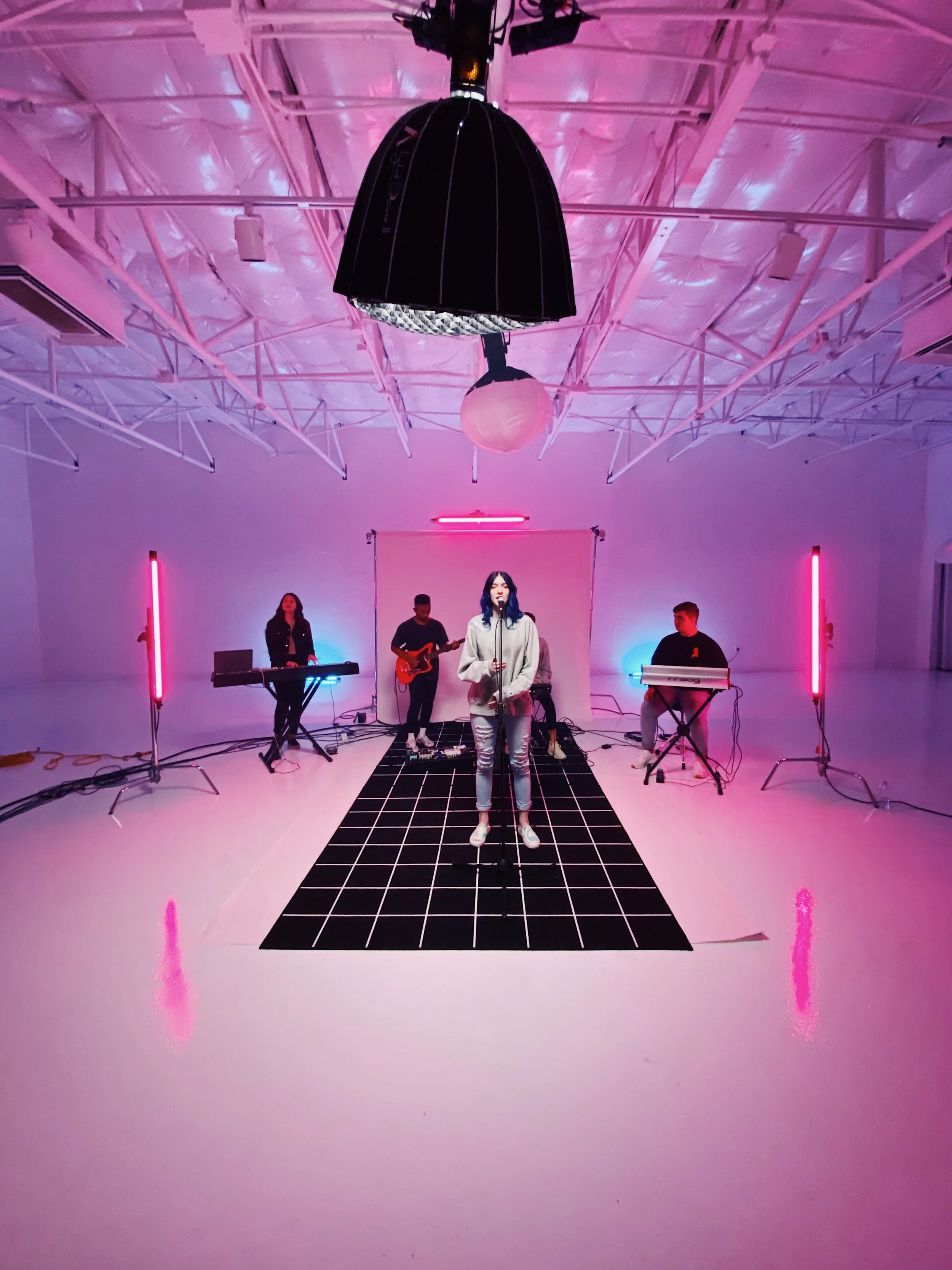 A band performing in a white studio with pink and blue lighting, featuring a female singer in front, and two keyboards and a guitar in the background.