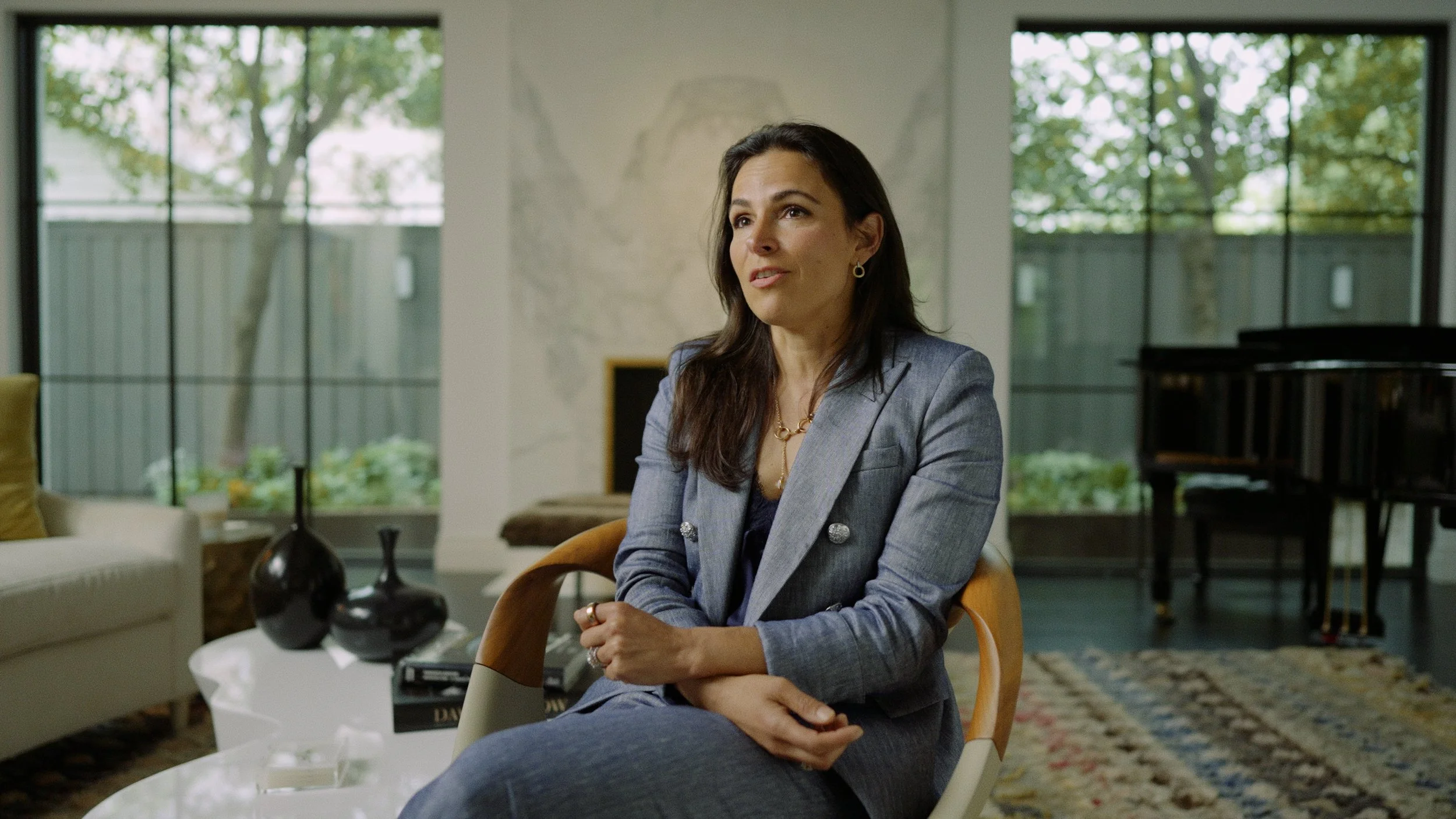 A woman with dark hair wearing a gray blazer and jewelry sitting in a modern living room with large windows, a piano, and a fireplace.