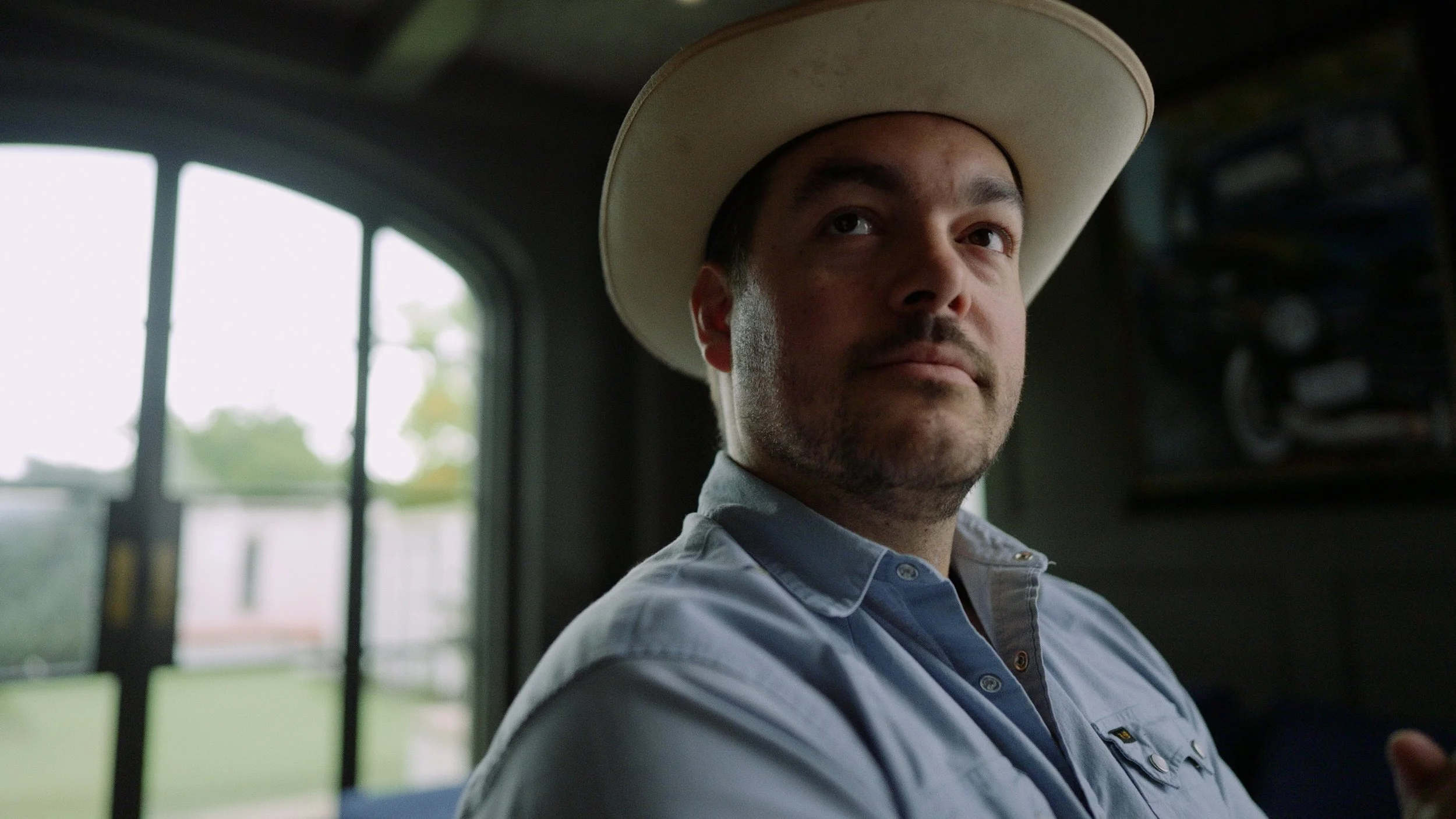 A man wearing a cowboy hat and a light blue button-up shirt looking to the side inside a room with a window showing a blurred outdoor scene.