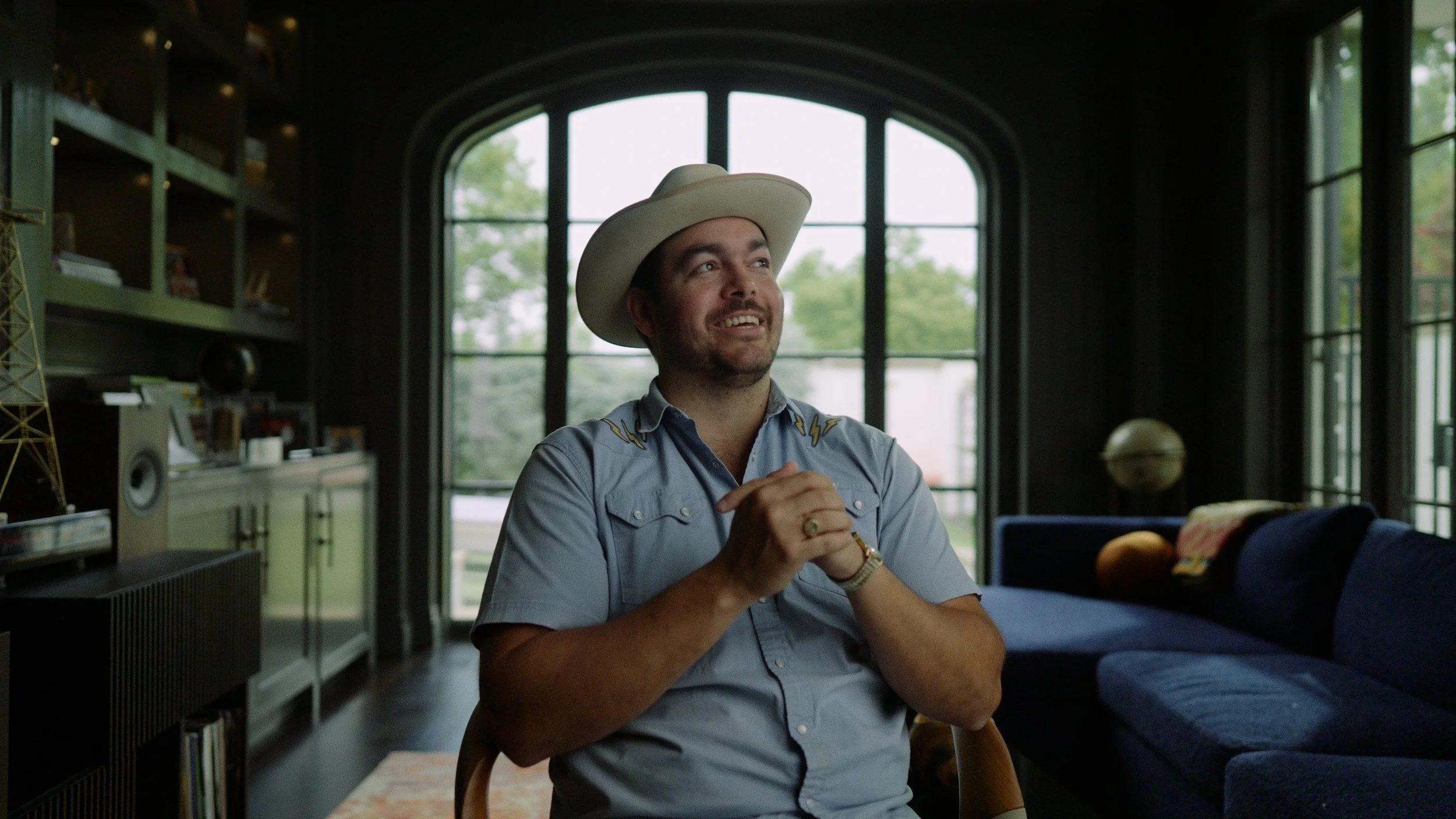 A man wearing a cowboy hat and a light blue shirt sits in a living room, smiling and looking up with clasped hands on his lap. The room has large windows, dark walls, and bookshelves.