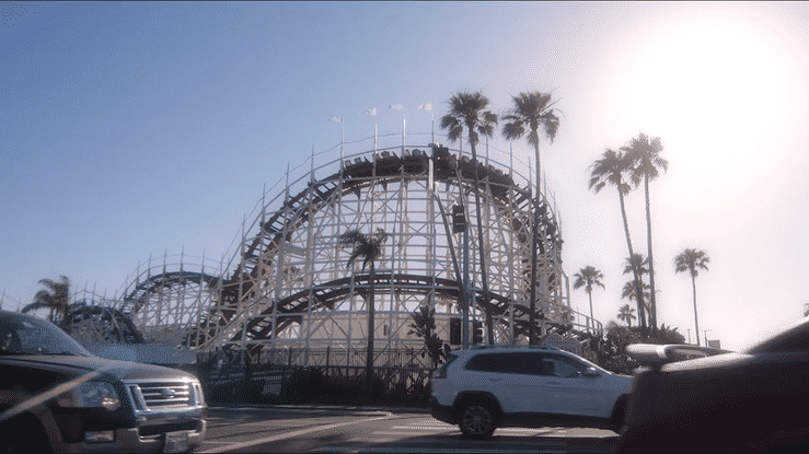 A wooden roller coaster with white supports and a black track at an amusement park, surrounded by tall palm trees, with parked cars in the foreground and a partly cloudy sky in the background.