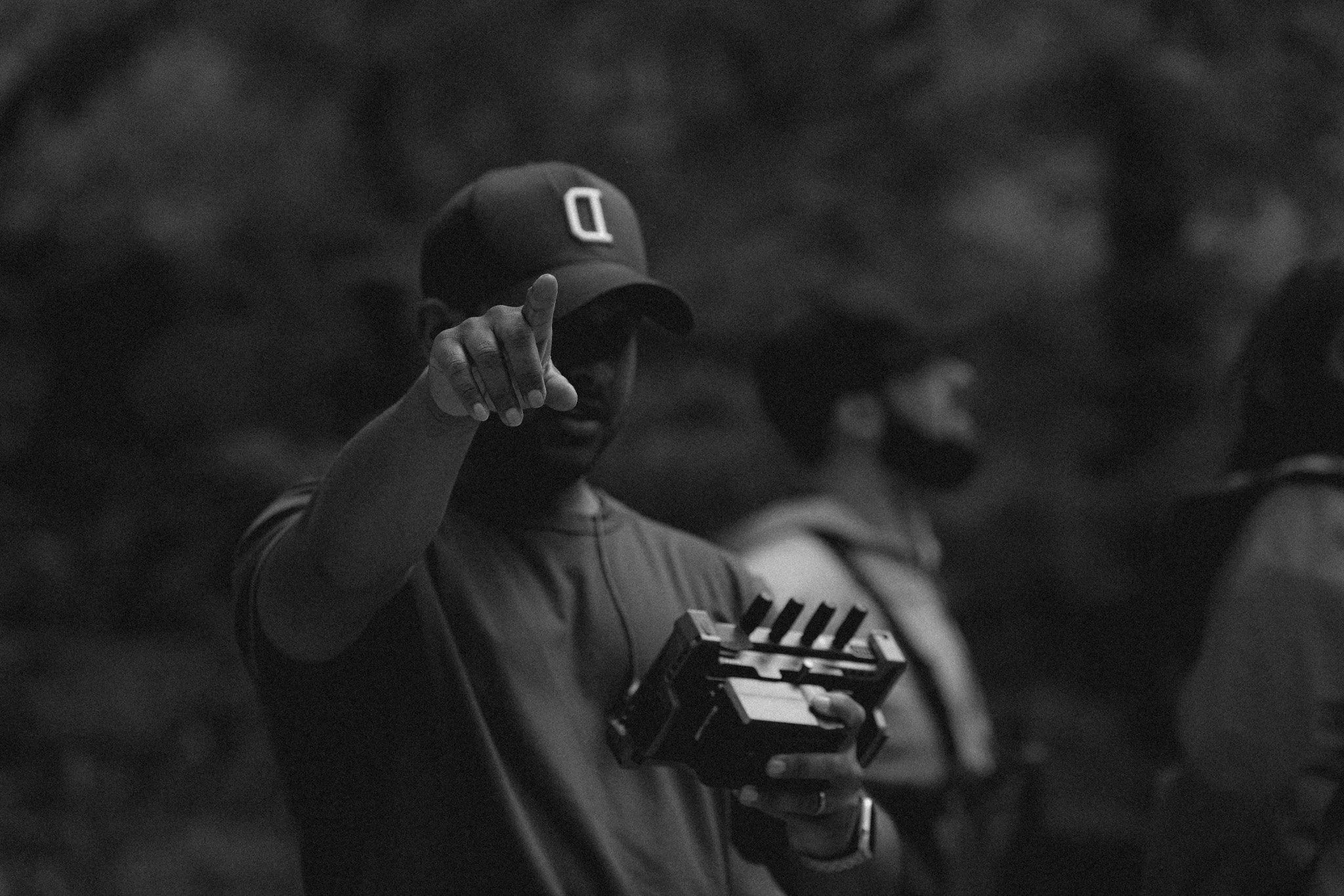 A person wearing a cap and face mask is pointing at the camera while holding a drone remote controller. Other people are blurred in the background.