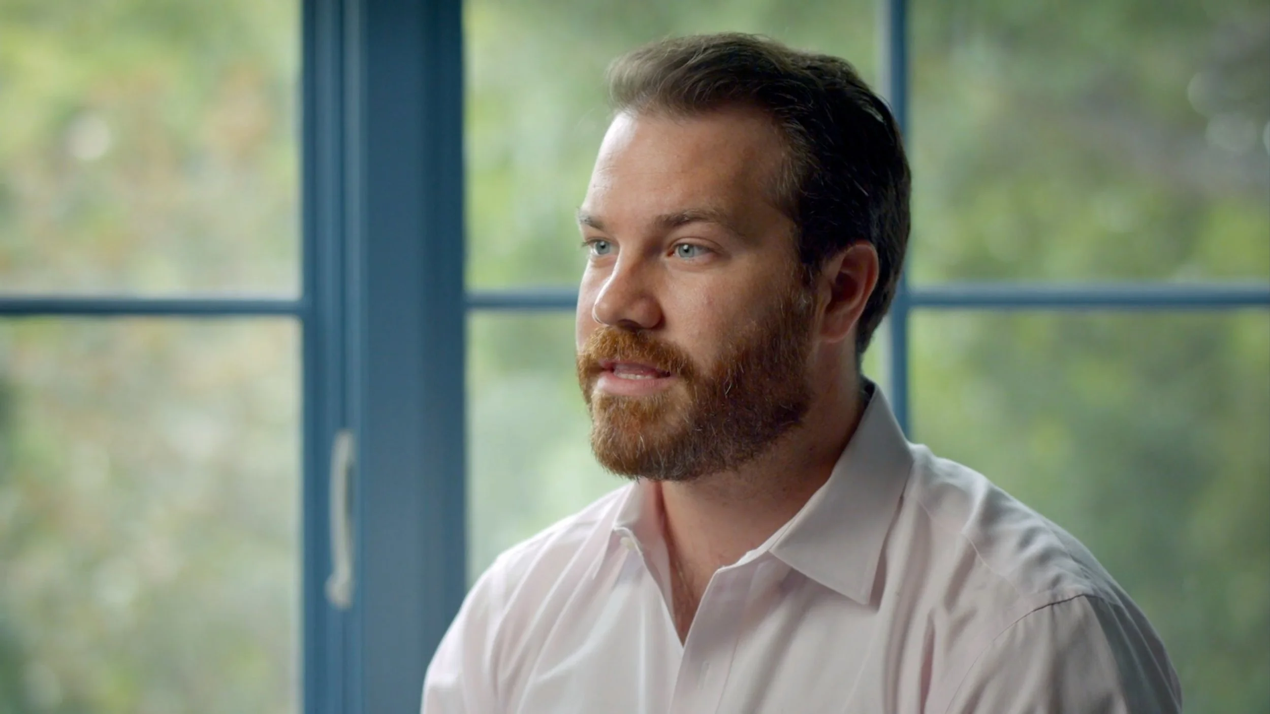 A man with a beard and blue eyes in a white button-down shirt sitting near a window with a blurred green outdoor background.