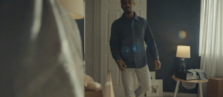 A man standing in a bedroom, looking at someone or something off-camera, with a bedside table, lamp, and curtains in the background.