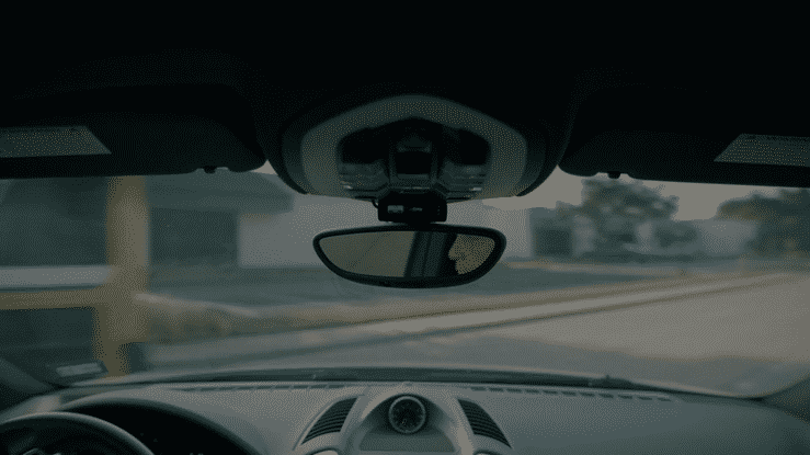 View from inside a car showing the dashboard and rearview mirror during daytime with a road ahead.