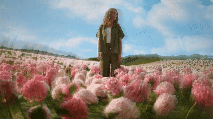 A woman with curly blonde hair wearing a green jacket and black pants standing in a field of pink and white flowers under a partly cloudy sky. Filmed in a XR studio. Virtual Production. LED wall.