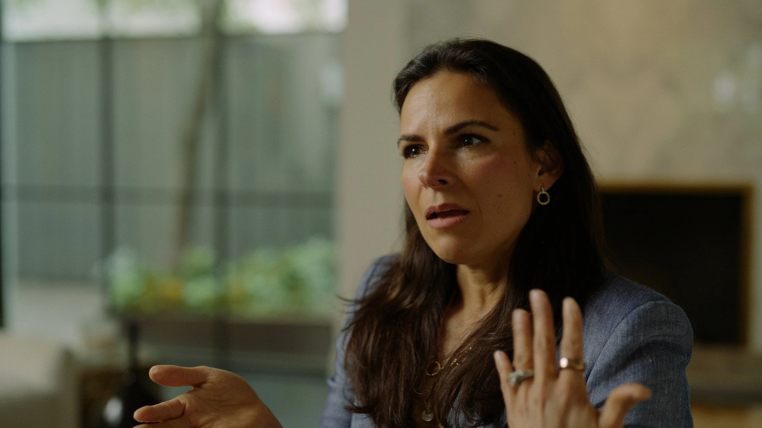 Close-up of a woman with dark brown hair, wearing earrings and a blazer, appearing to be speaking or explaining something with a serious or concerned expression, indoors with blurred background.