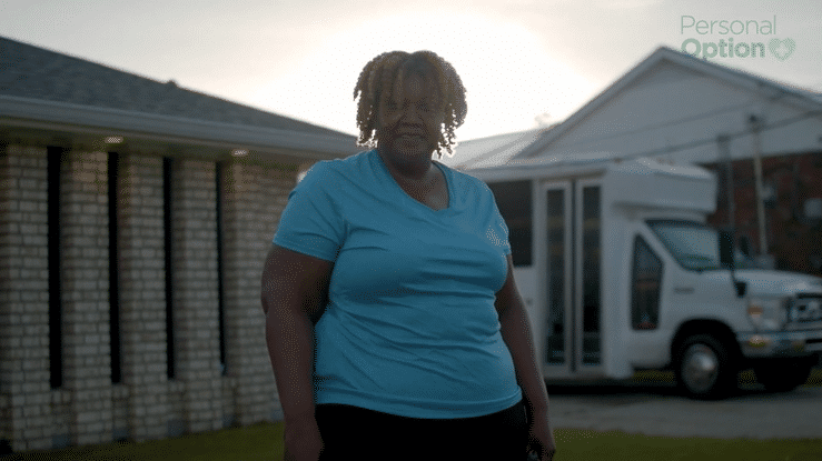 A woman stands outdoors in front of a brick building and a truck, wearing a blue t-shirt, with the sun behind her.