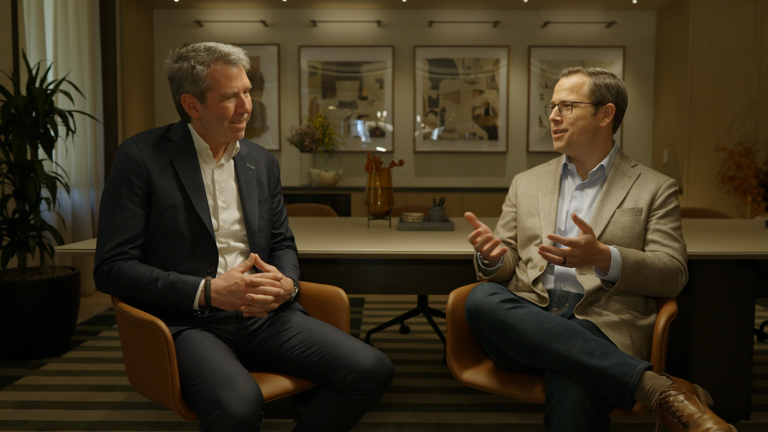 Two men having a conversation in an office setting. One wears a black blazer and white shirt, the other a beige blazer and light blue shirt with glasses.