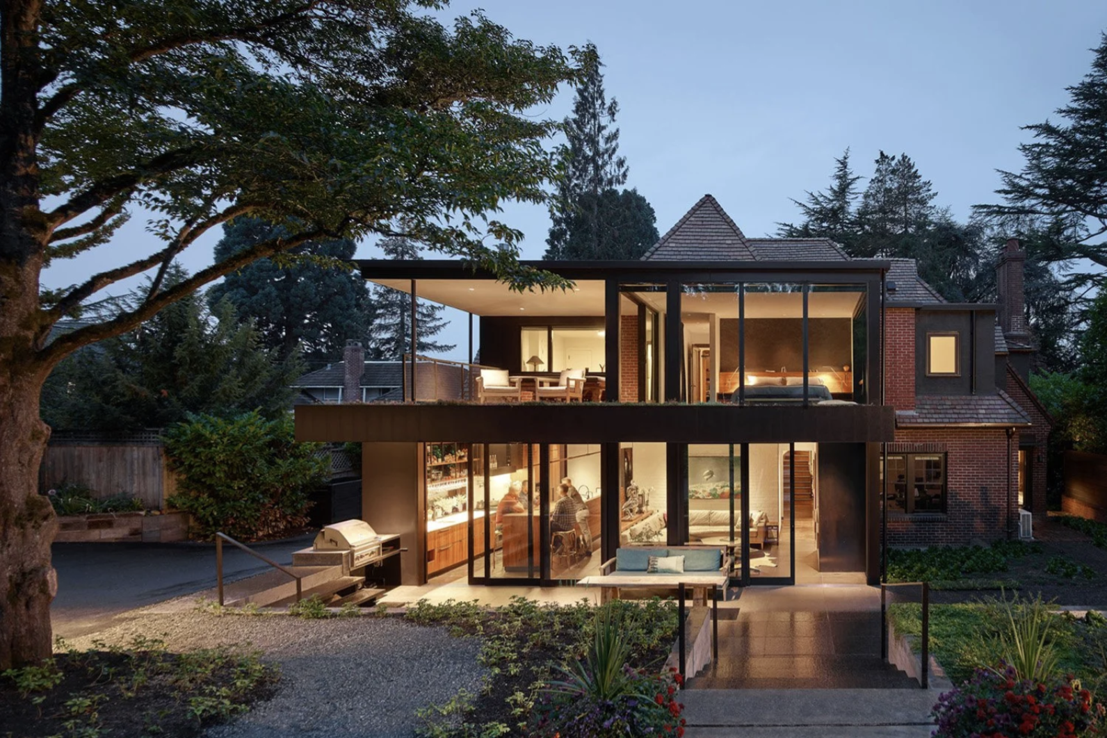 Modern backyard addition of a French Norman style house with walls of glass lit from within at dusk.
