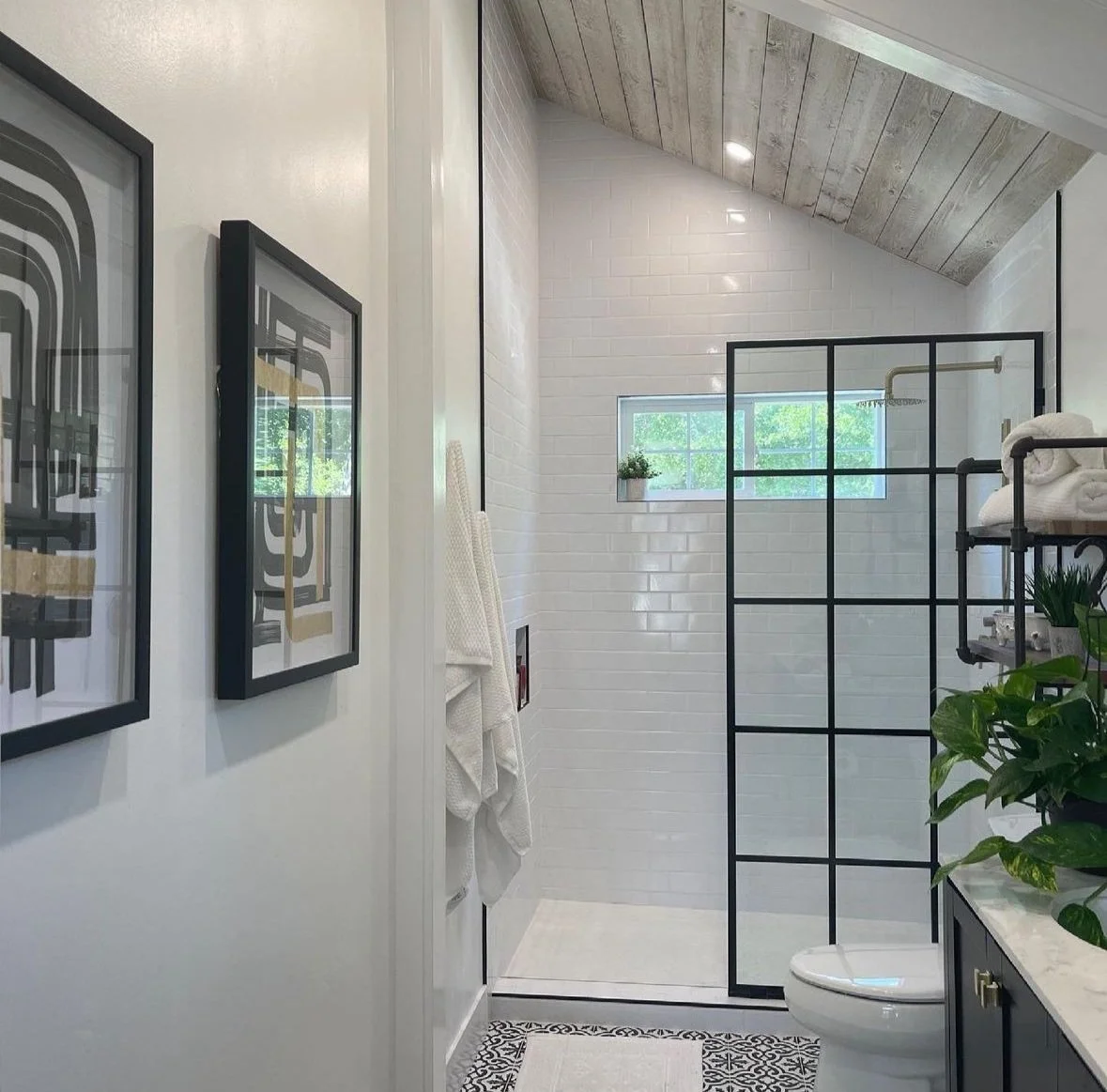Modern bathroom with white tiled walls, a rain shower, small window, black-framed glass partition, and decorative plants.