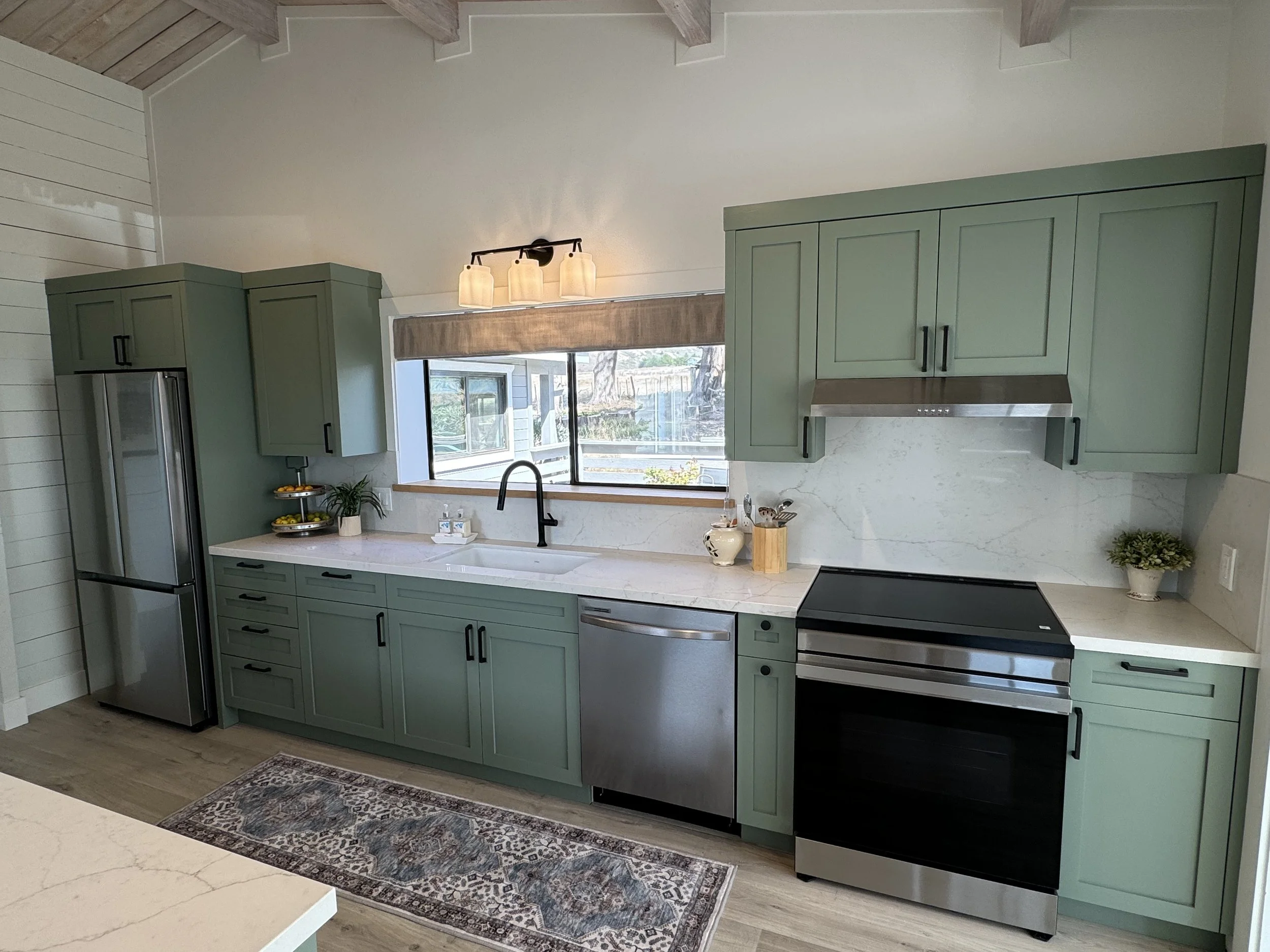 Modern kitchen with sage green cabinets, stainless steel appliances, a white marble backsplash, and a large window above the sink letting in natural light.