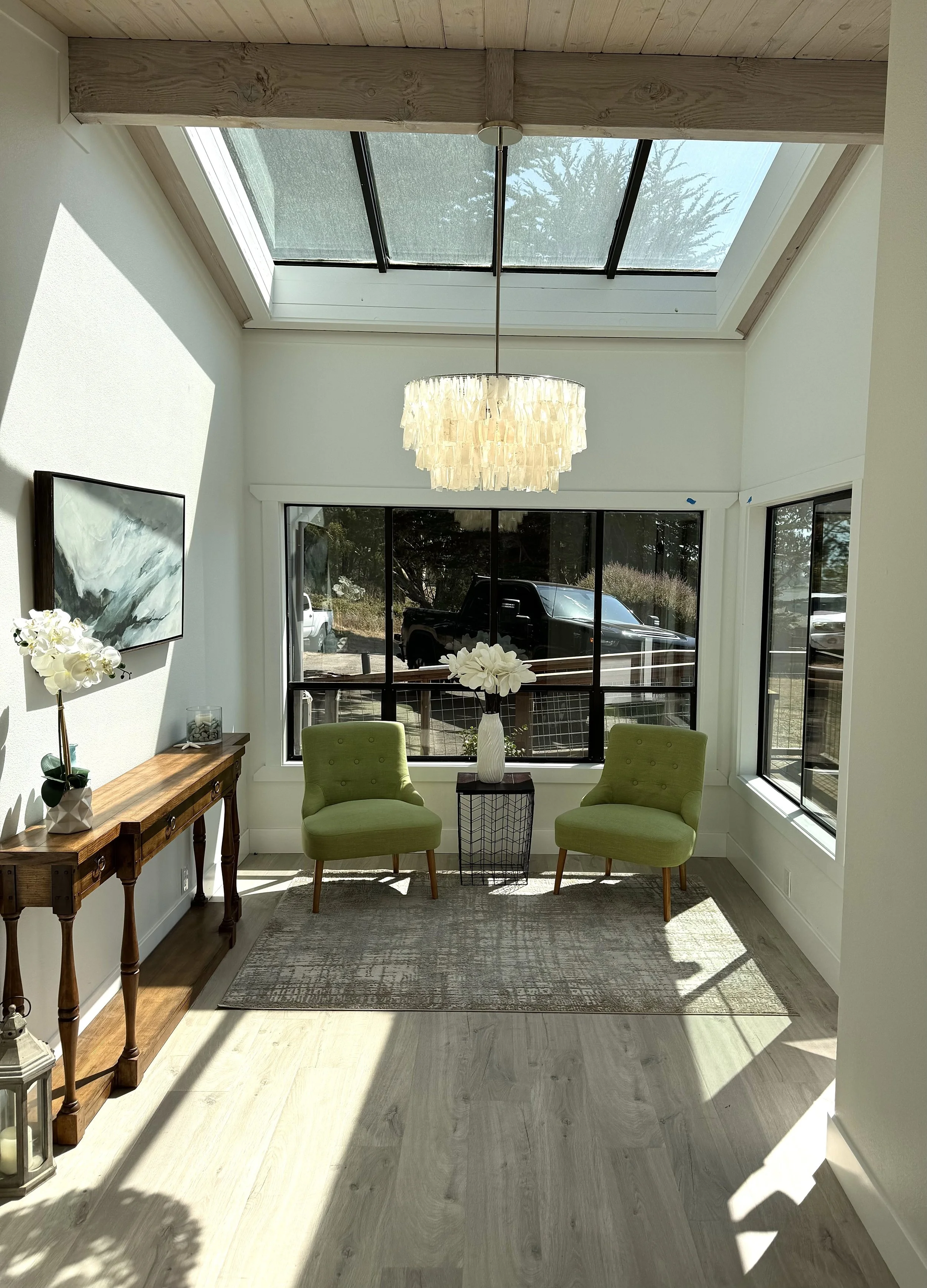 Sunlit room with two green chairs, a vase with white flowers between them, a wooden console table with flower and candle decor, a hanging chandelier, large window, and skylight, with a view of trees and a black truck outside.