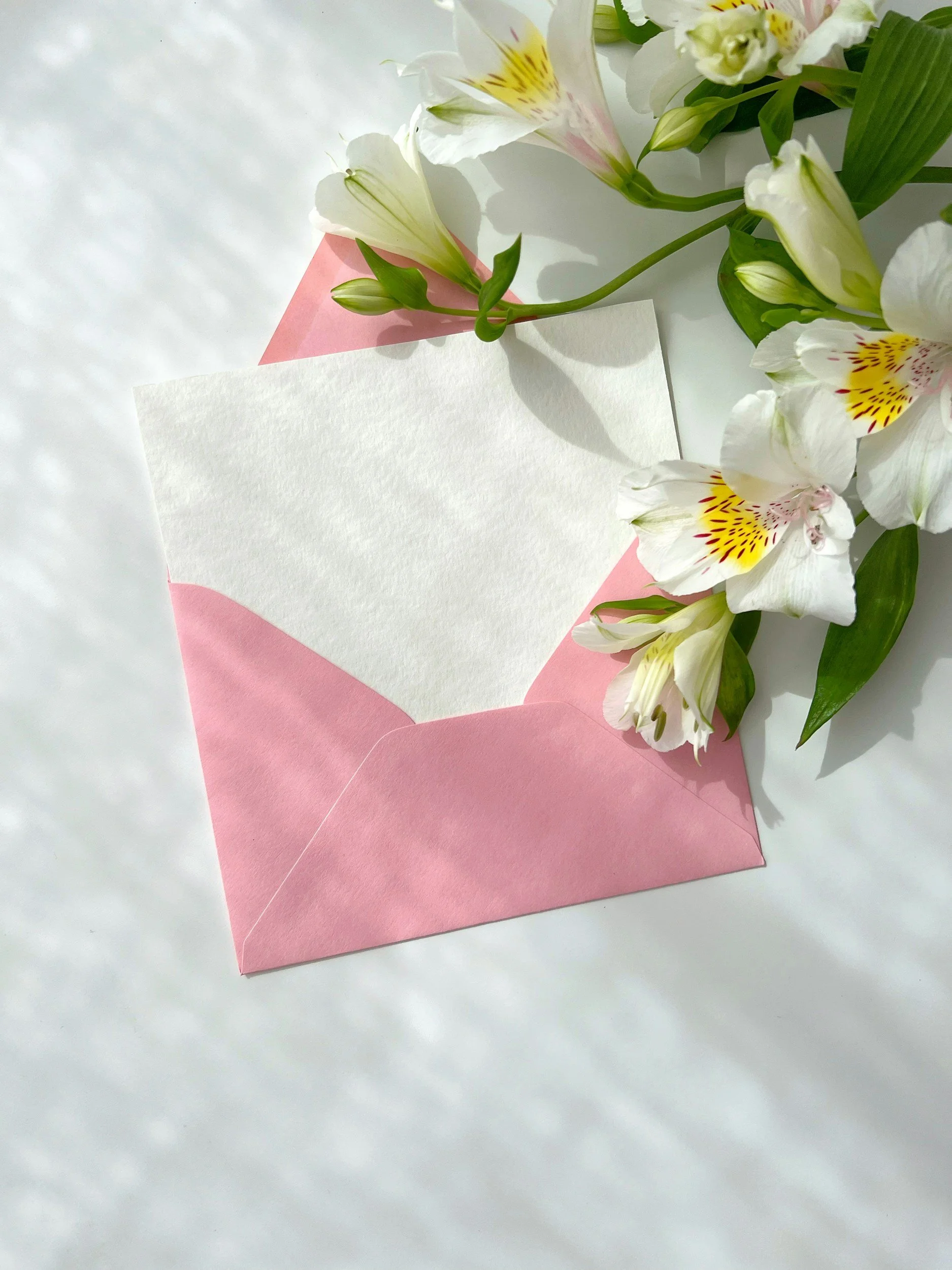A pink envelope with a blank white letter inside, surrounded by white and pink flowers, on a white surface.