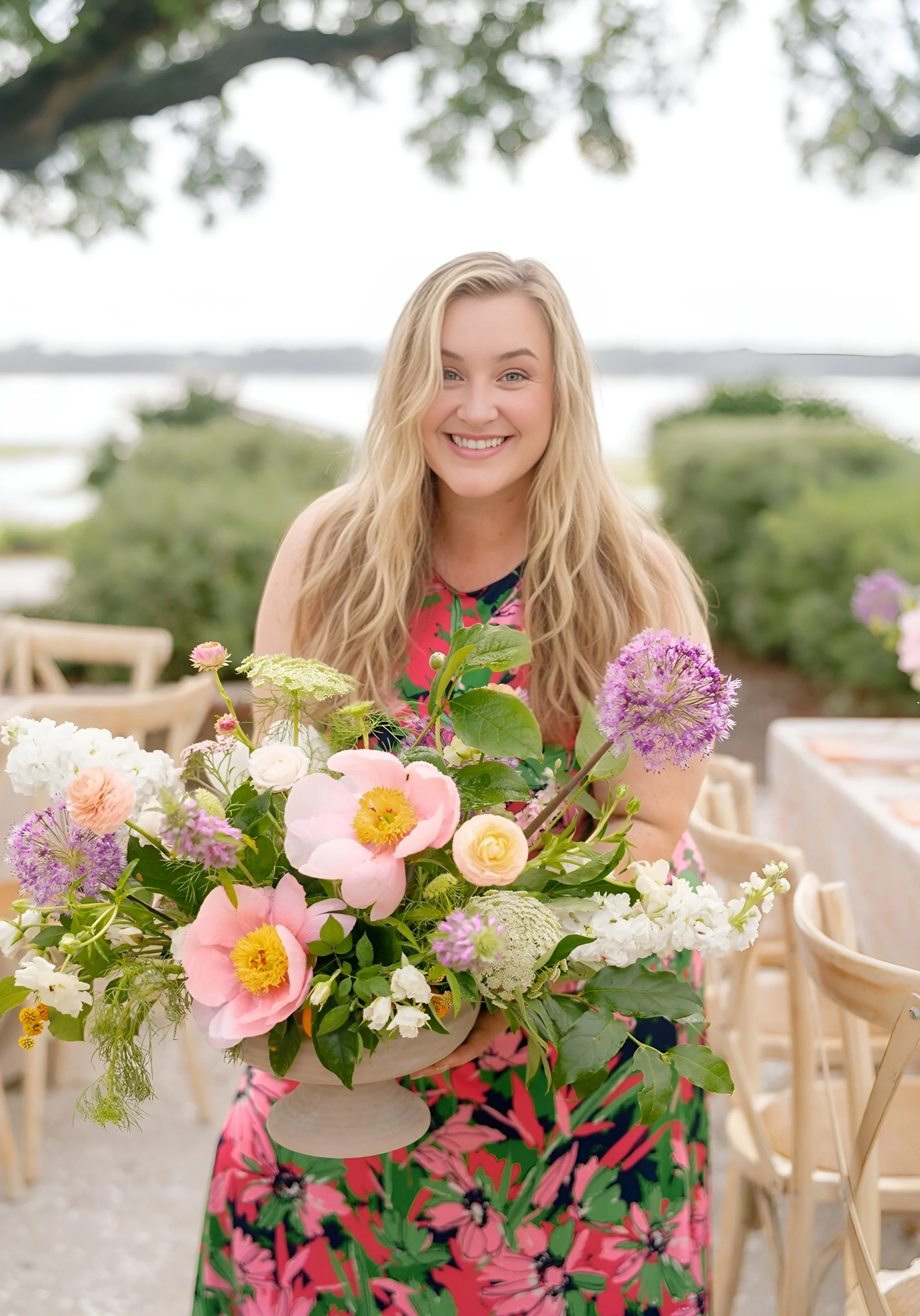 A smiling woman in a colorful floral dress holding a large bouquet of pink, white, and purple flowers outdoors near water and greenery.