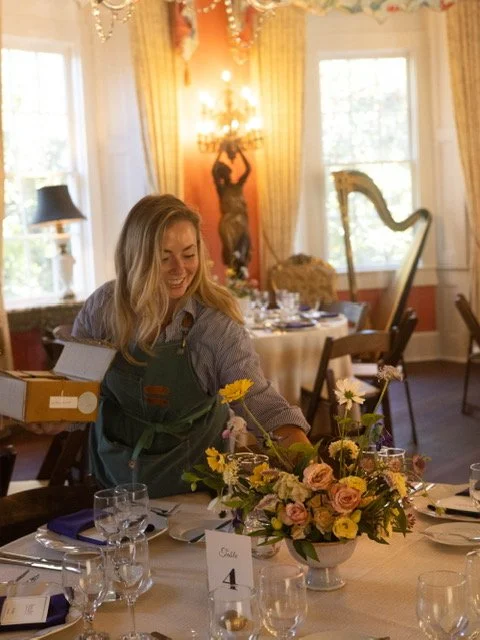 A woman setting a dining table with a floral centerpiece in an elegant room with natural light, golden curtains, and artistic decor.