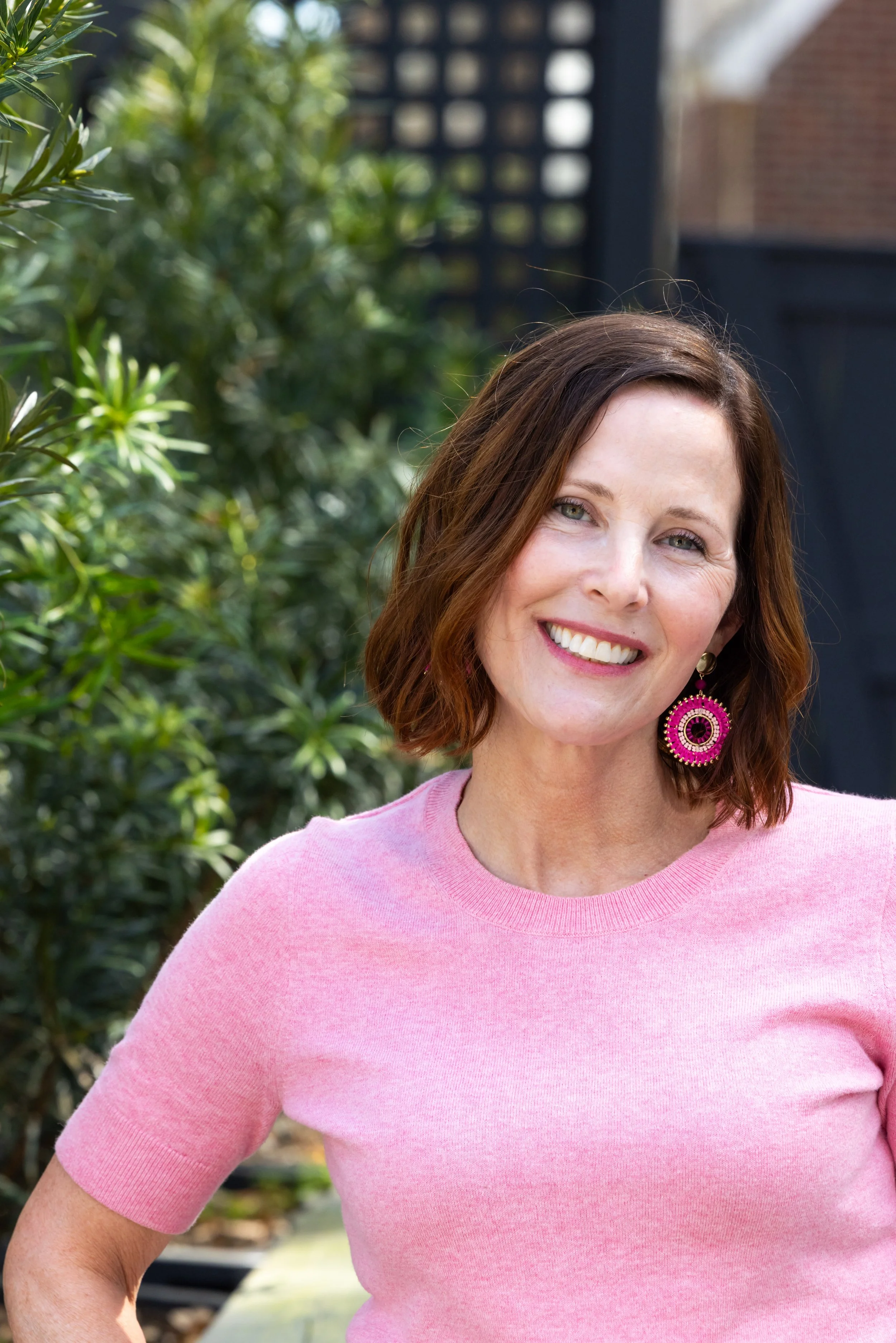 A woman with shoulder-length brown hair smiling outdoors in front of greenery, wearing a pink short-sleeve shirt and pink circular earrings.