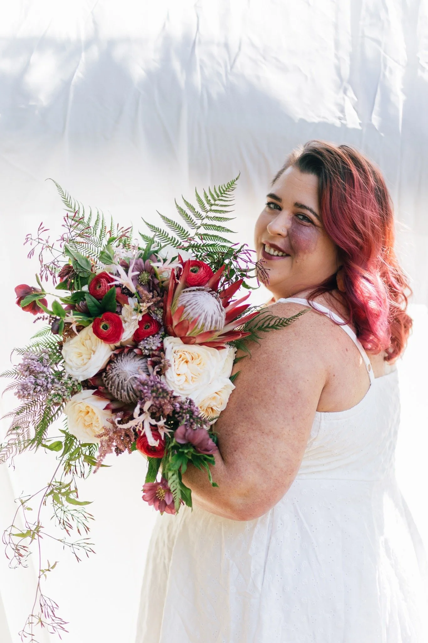 Bridal portrait of a woman with pink hair holding a large bouquet of colorful flowers, wearing a white wedding dress.