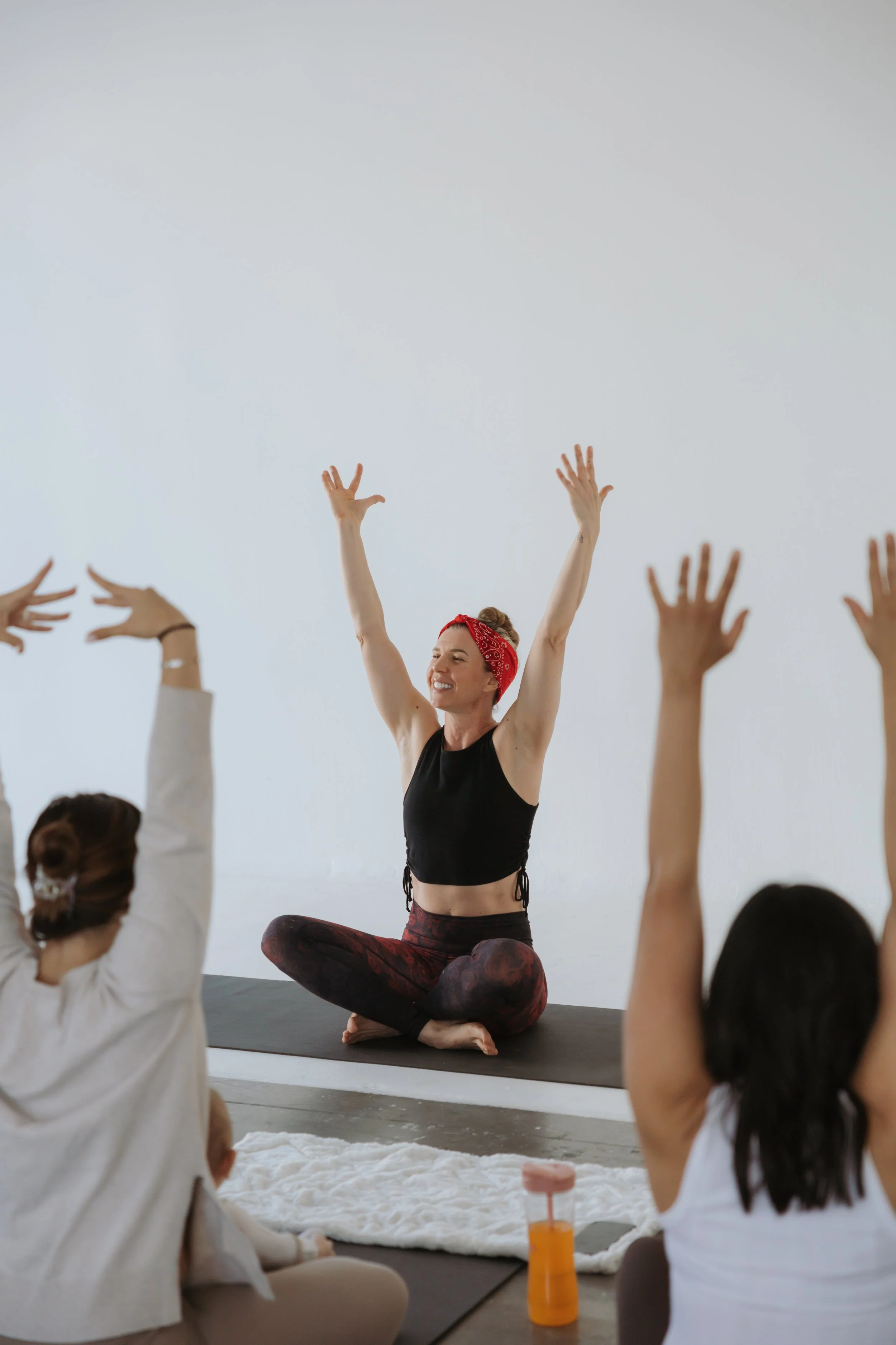 A woman leading a yoga class, seated on a yoga mat with her arms raised, smiling, surrounded by others in a yoga studio.