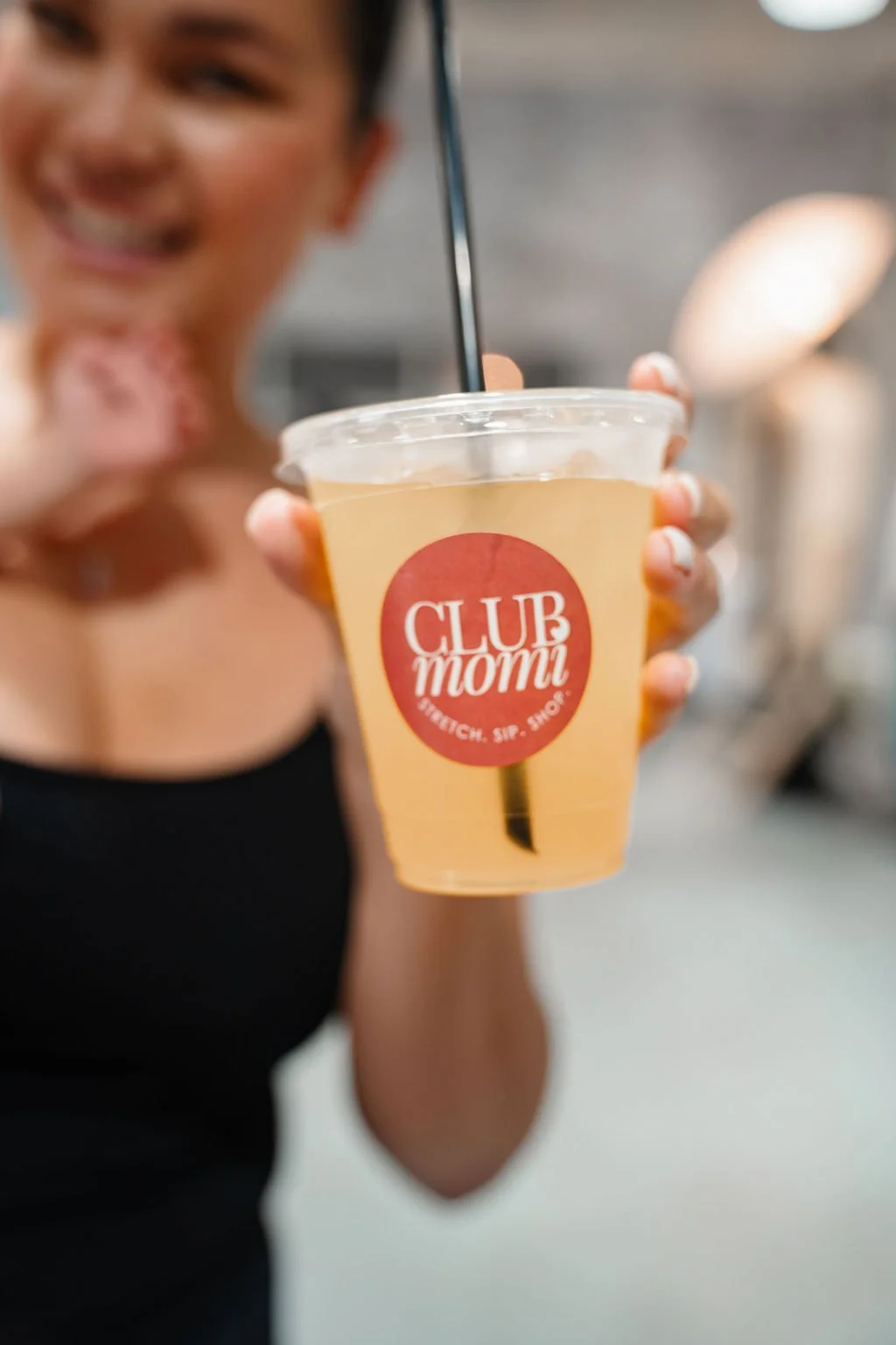 Smiling woman holding a clear plastic cup of yellow beverage with a red and white "CLUB mom" logo and a black straw, in an indoor setting.