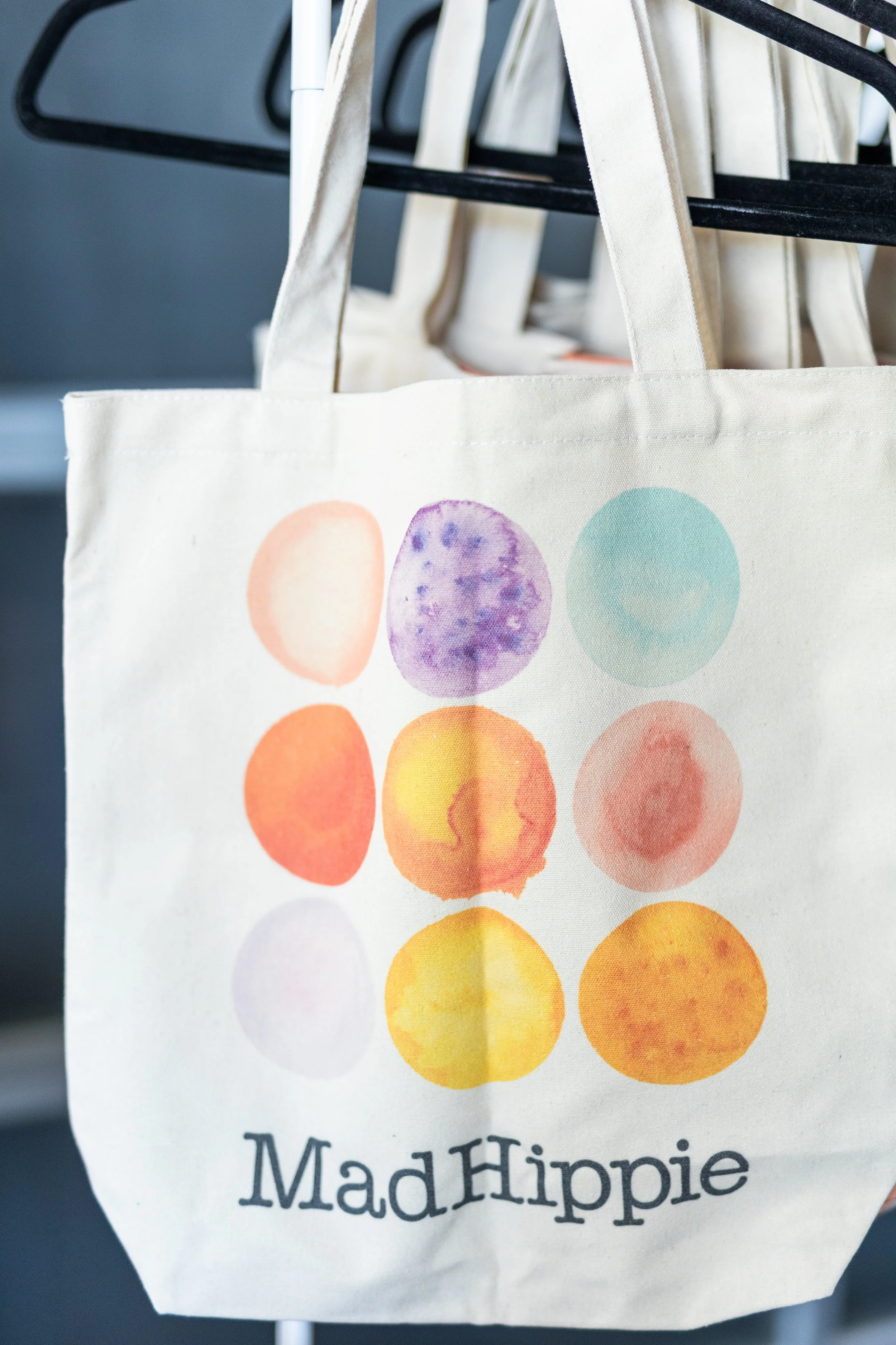 A beige tote bag hanging on a black rack, decorated with watercolor-style circles in pastel colors and the text "Mad Hippie" at the bottom.