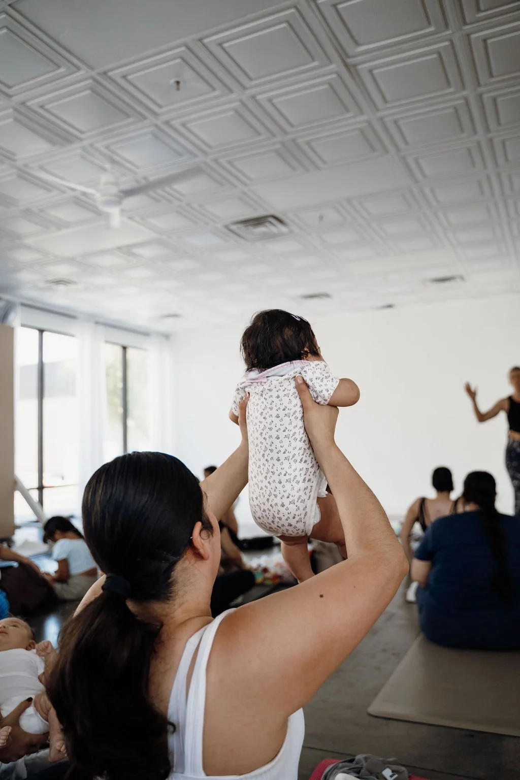 A woman holding up a small child in a room with other people. The room has large windows and a decorated ceiling. There is a woman at the front using expressive hand gestures.