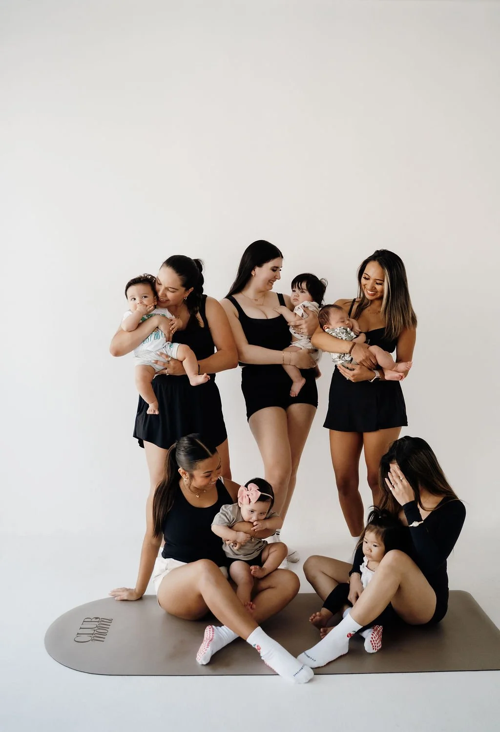 Six women sitting and standing with multiple babies, all wearing black outfits, in front of a plain white background.
