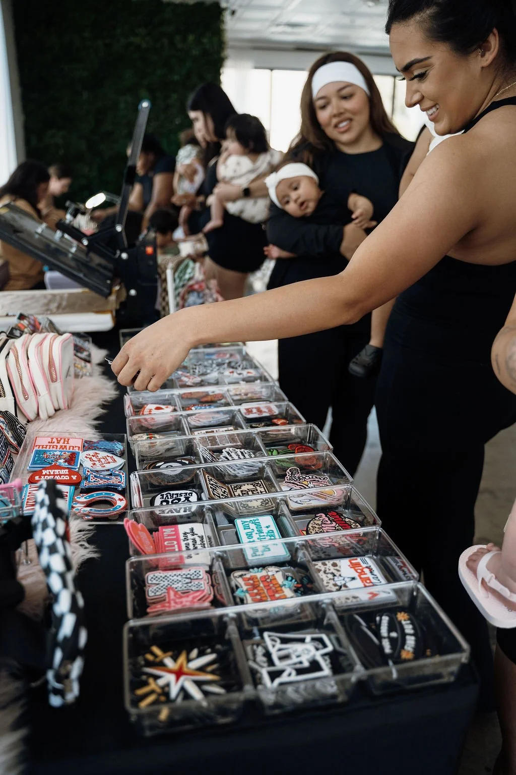 Women shopping for patches and pins at a table in a crowded indoor market or fair.