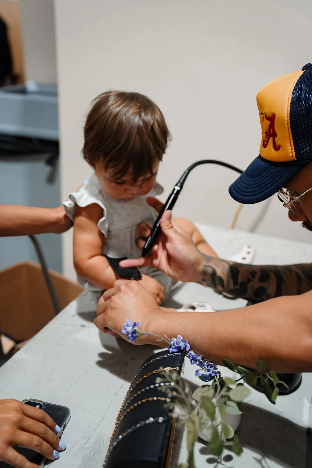A young child sitting on a table being examined by a man wearing a blue Alabama hat and glasses. The man is holding the child's chest area with one hand and a stethoscope with the other, while the child looks down at the stethoscope. A woman with a t