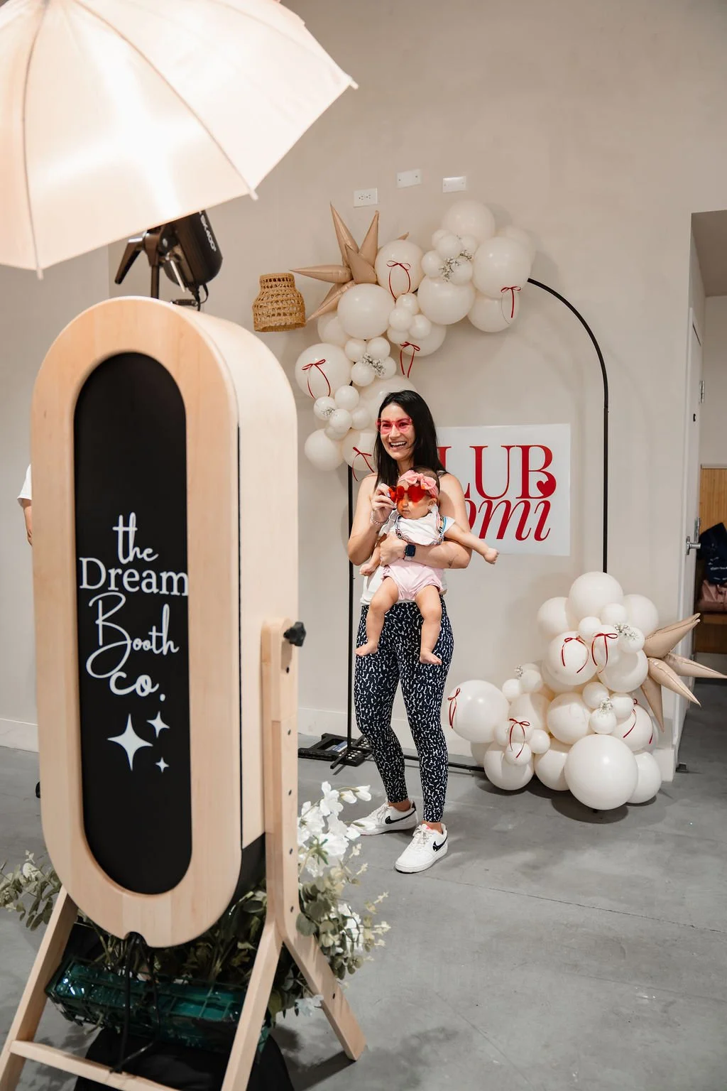 A smiling woman holding a young girl in front of a balloon arch and a sign at an indoor event. The woman wears pink sunglasses and athletic shoes, while the girl has a flower headband and oversized sunglasses.