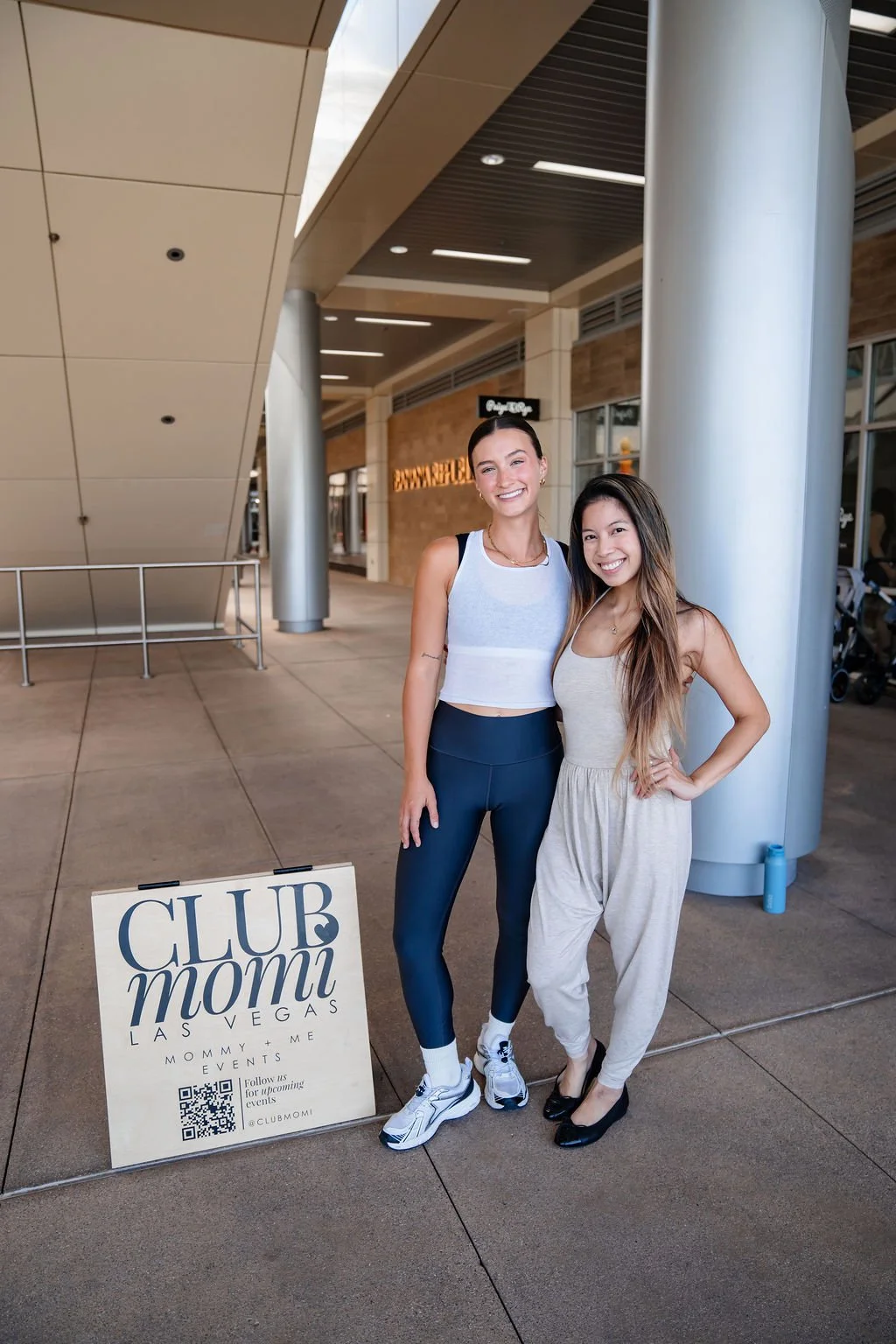 Two women standing outside a shopping mall, smiling at the camera. One is wearing athletic clothing, and a sign in front of them reads 'Club Mommi Las Vegas'.